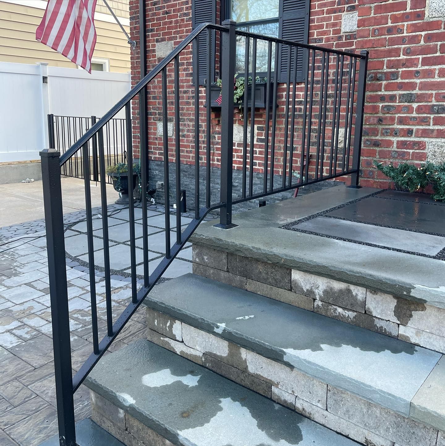 Black metal handrail on stone steps leading to a brick building entrance, with an American flag visible in the background.
