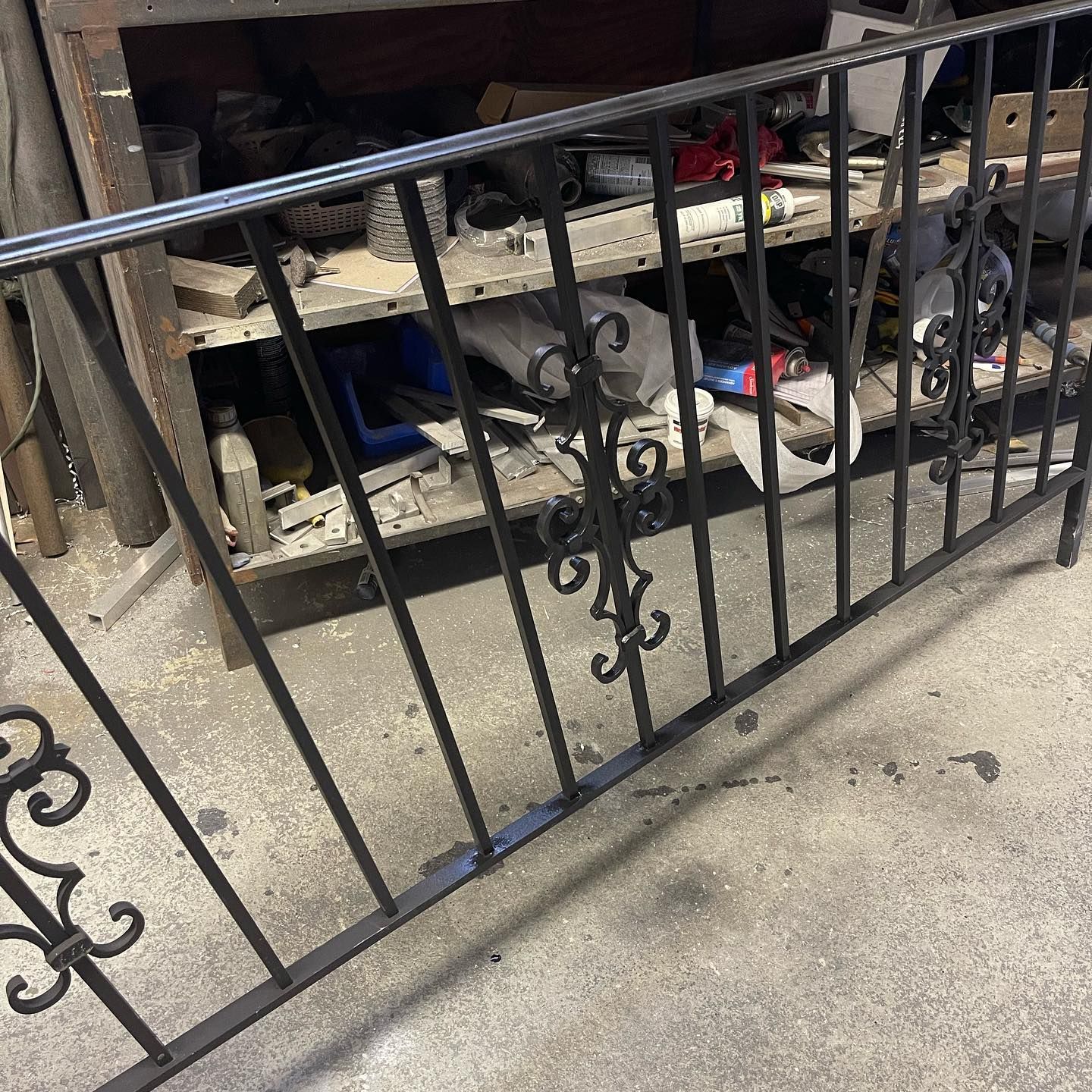 A black metal railing with ornate, scrolled scrollwork panels, standing on a concrete floor in a workshop.