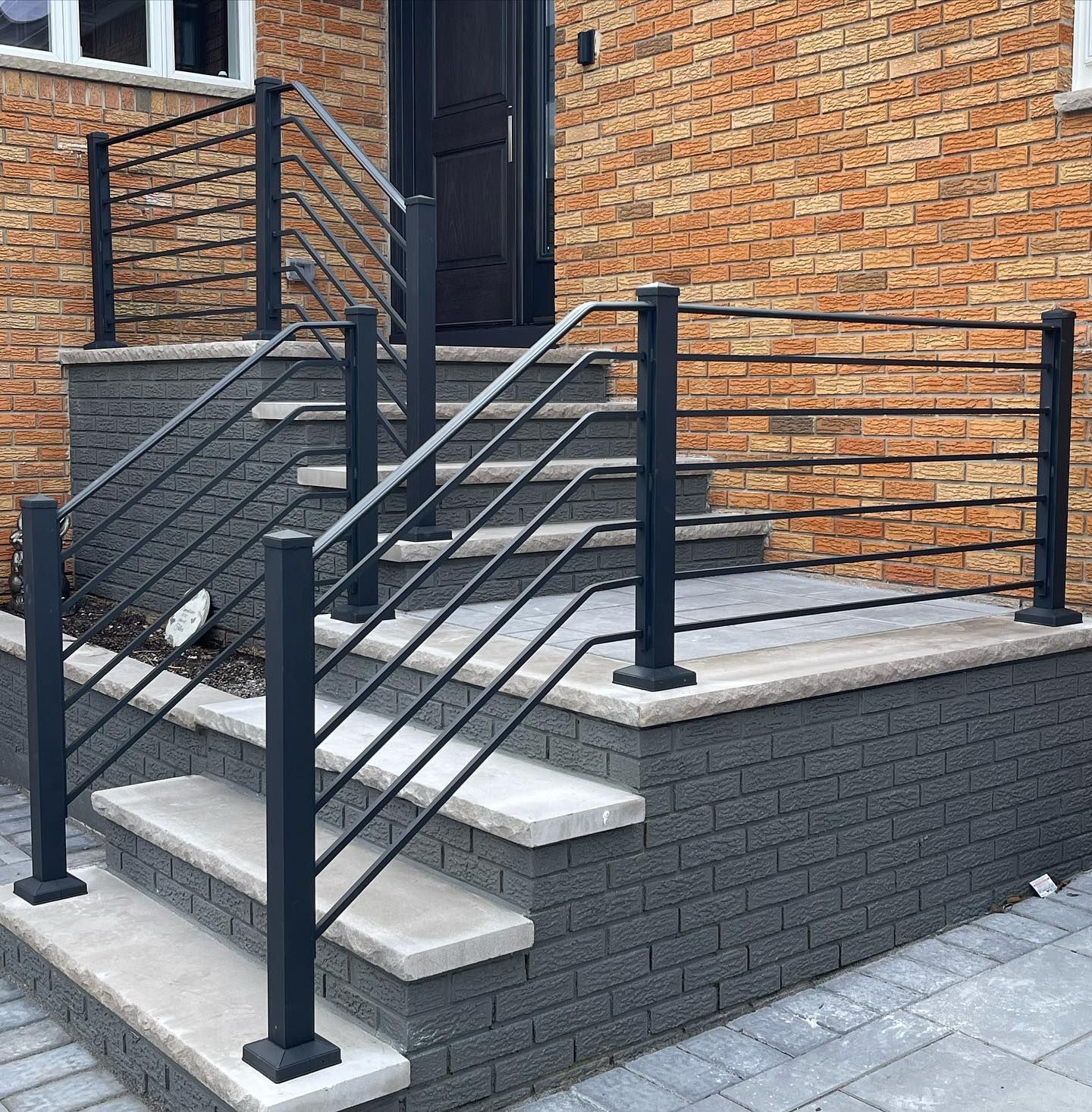 Black metal railings lining gray concrete steps leading to a dark door against a tan brick house exterior.