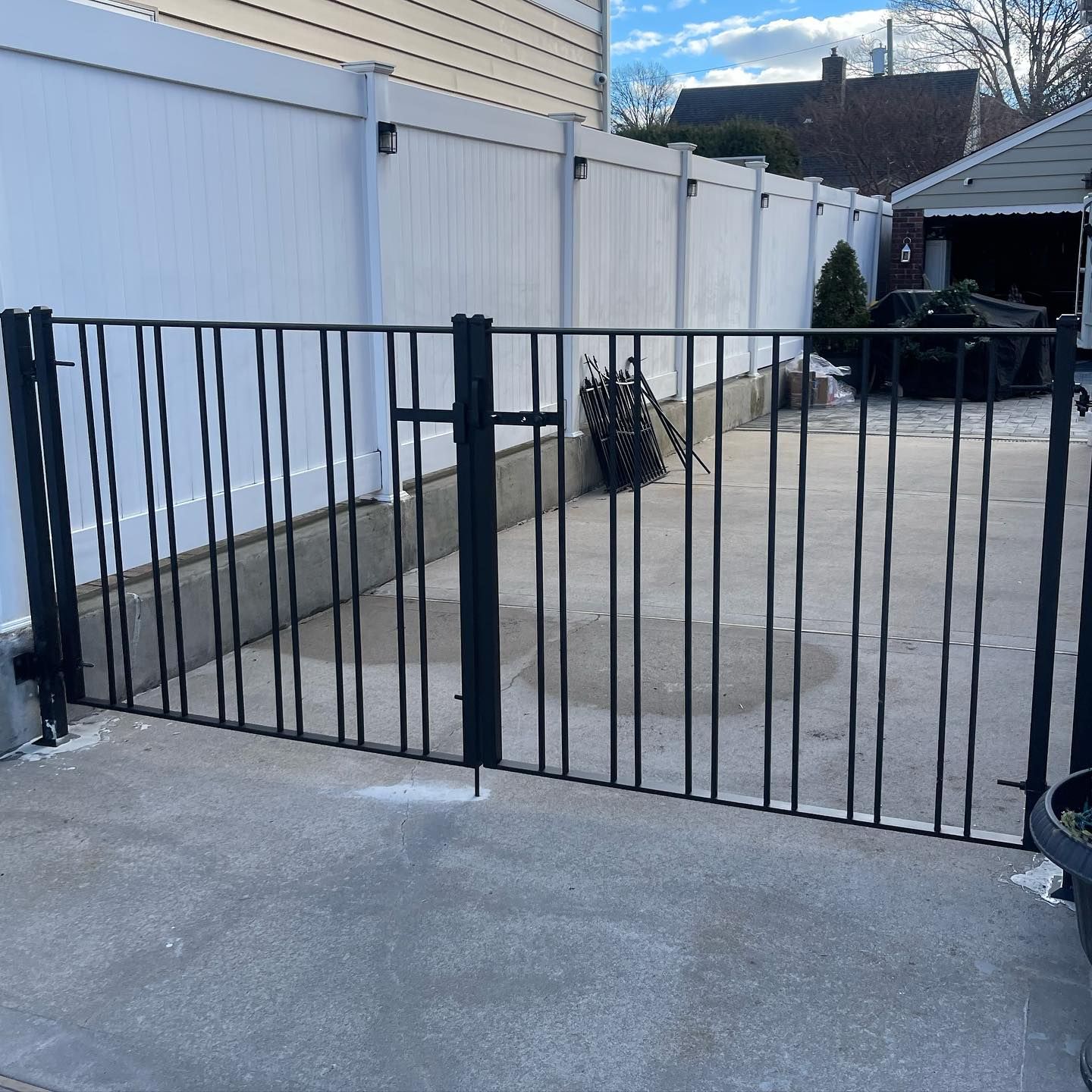A black metal gate stands in a driveway in front of a white vinyl fence and a house under a partly cloudy sky.
