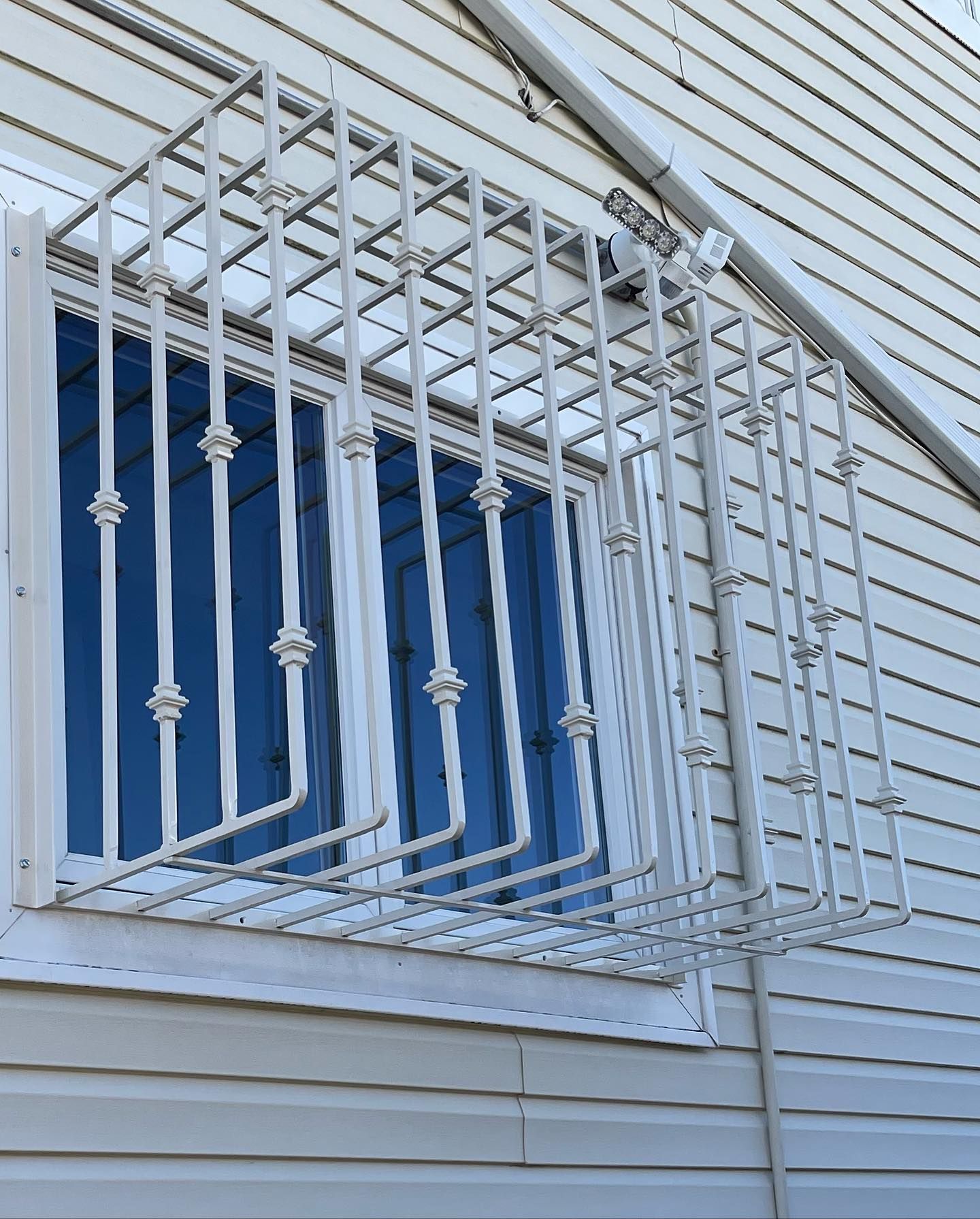 A white metal security cage mounted over a window on the exterior of a building with white siding.
