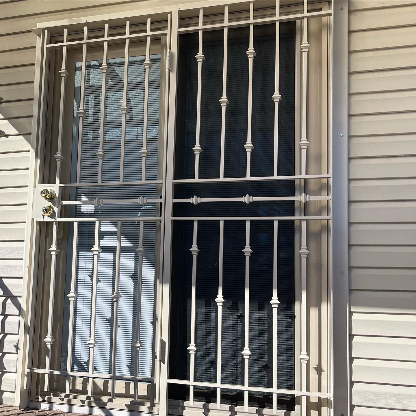 A cream-colored metal security door with vertical bars and decorative accents, installed on a house with beige siding.