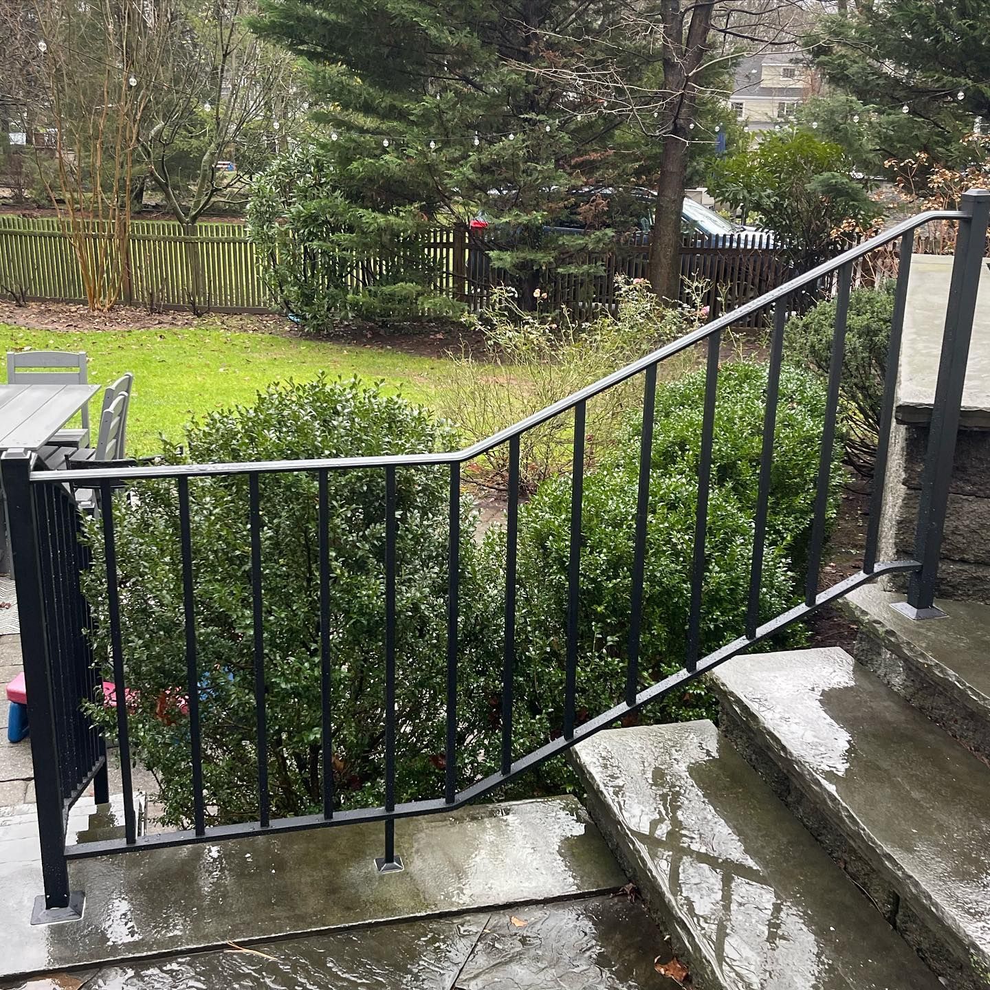 Black metal handrail on stone stairs leading to a green backyard with trees and a wooden fence.