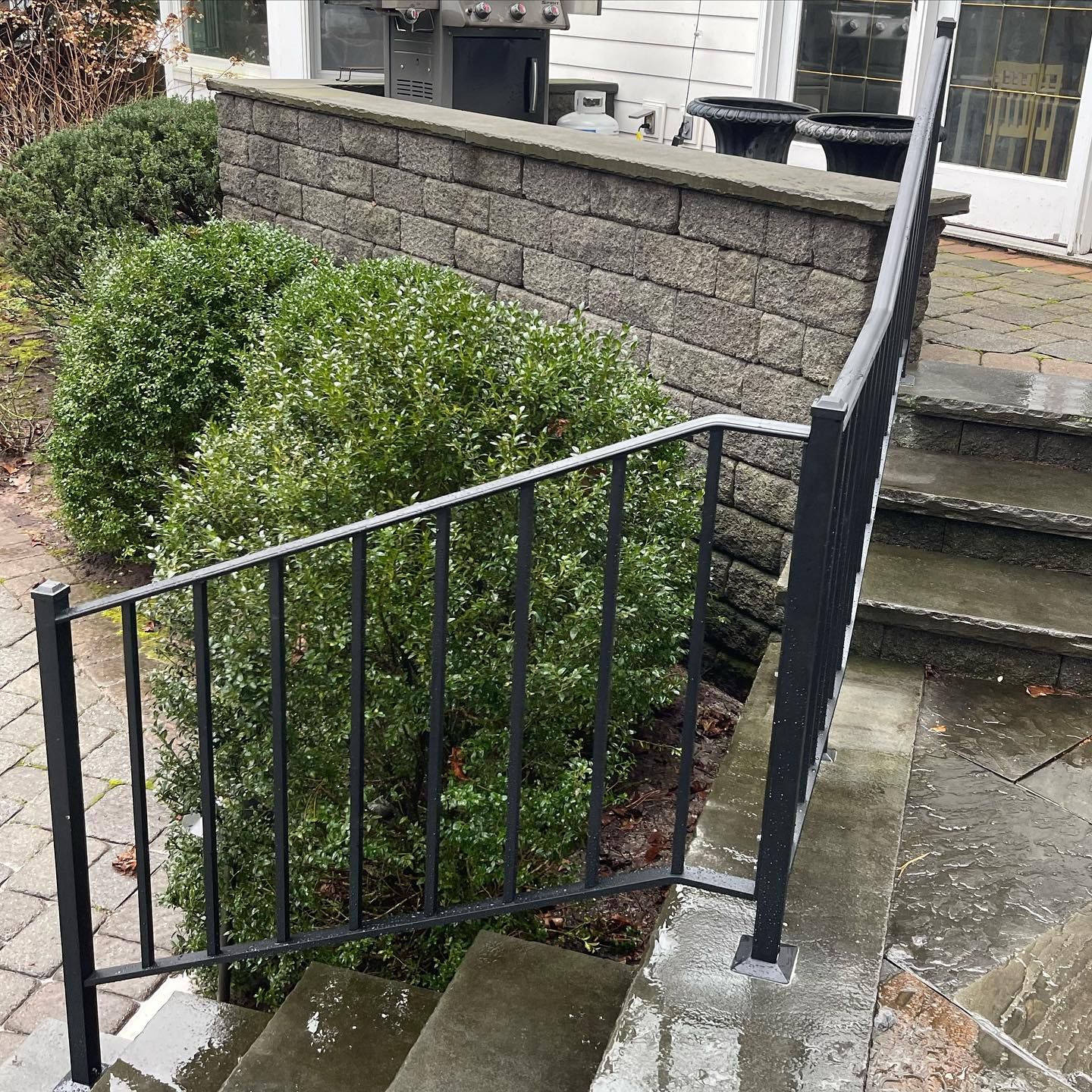Black metal handrail running alongside concrete outdoor stairs next to a brick wall and a large, rounded green shrub.