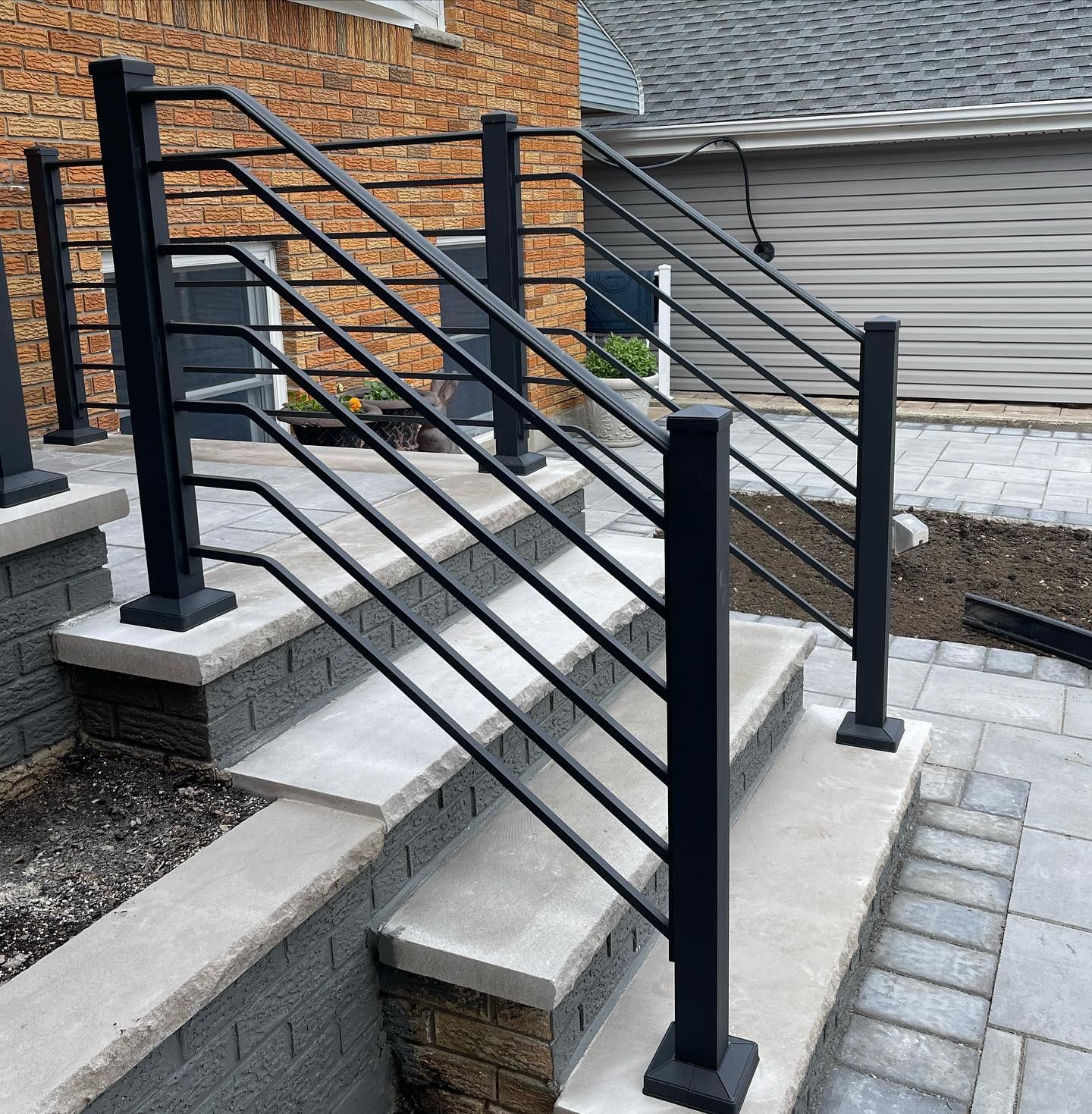 Black metal horizontal railing installed on outdoor stone steps leading to a brick building.