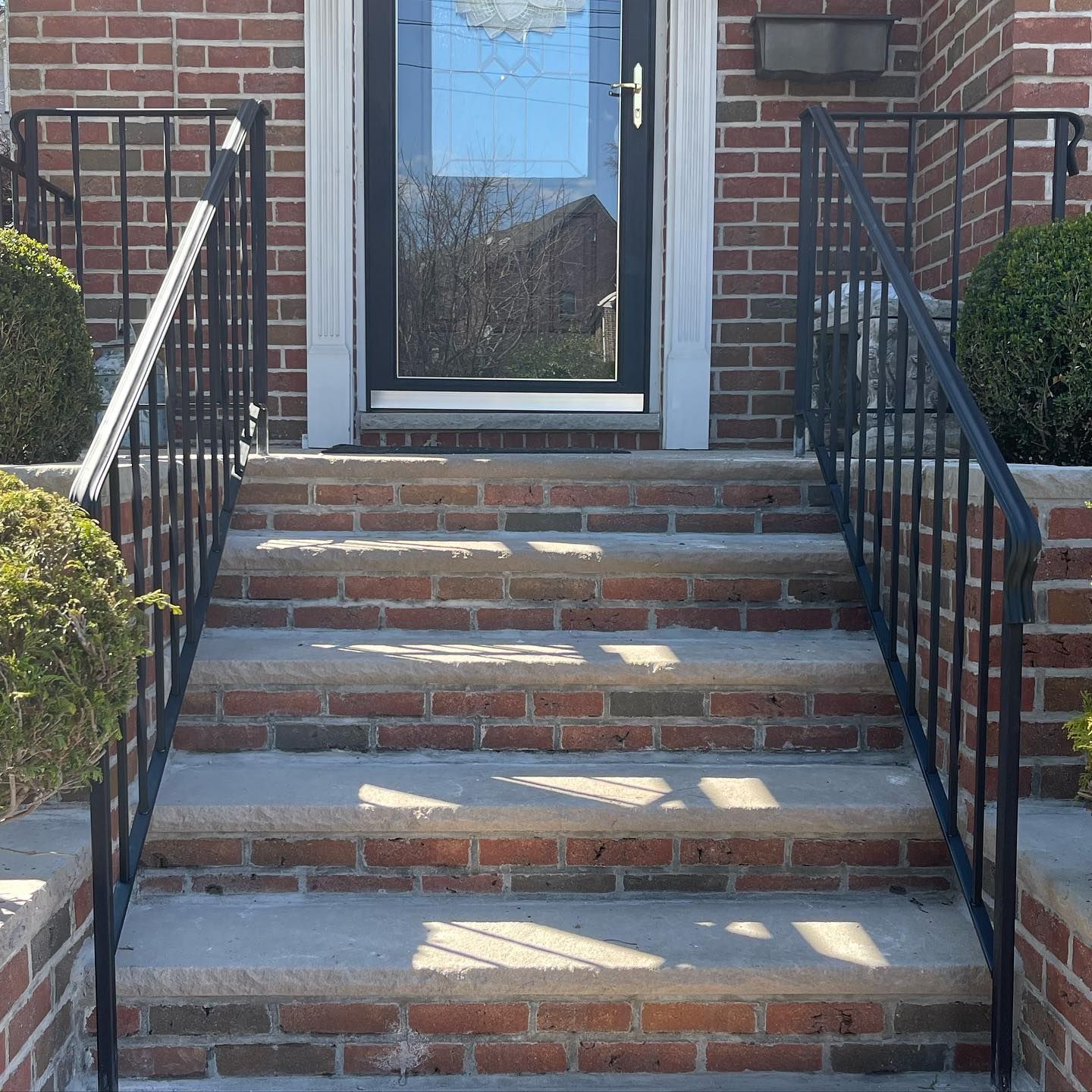 Brick steps leading to a dark glass front door with black metal railings on both sides and round shrubs.