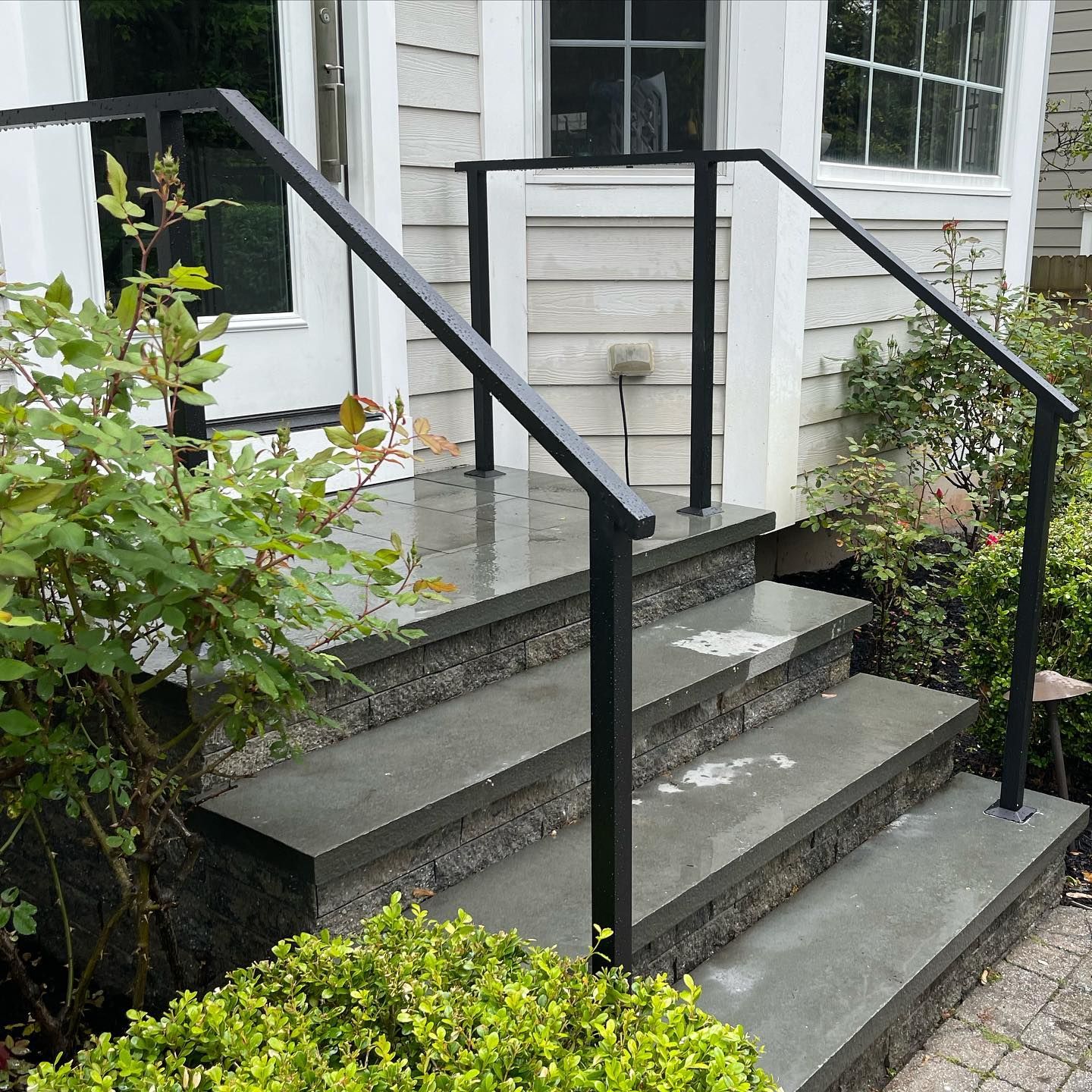 A set of three concrete steps leading to a house entrance, featuring black metal railings on both sides.