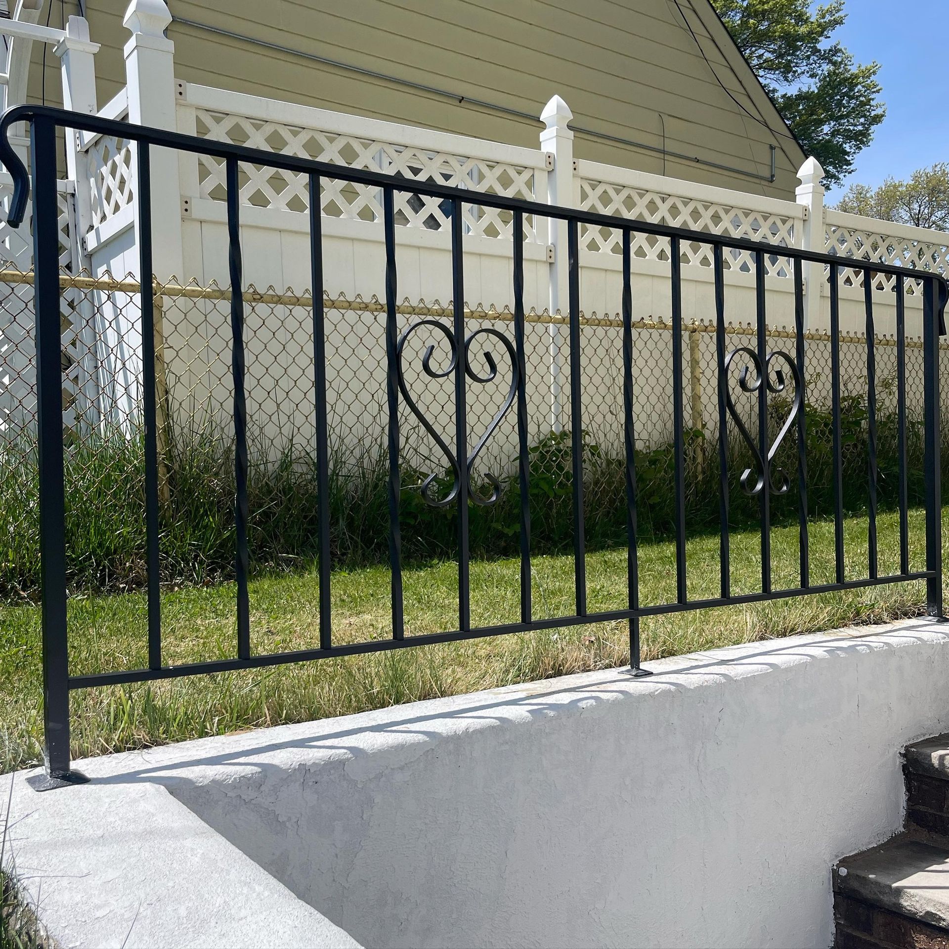 A black metal railing with heart-shaped decorative accents sits atop a concrete ledge in front of a white lattice fence.