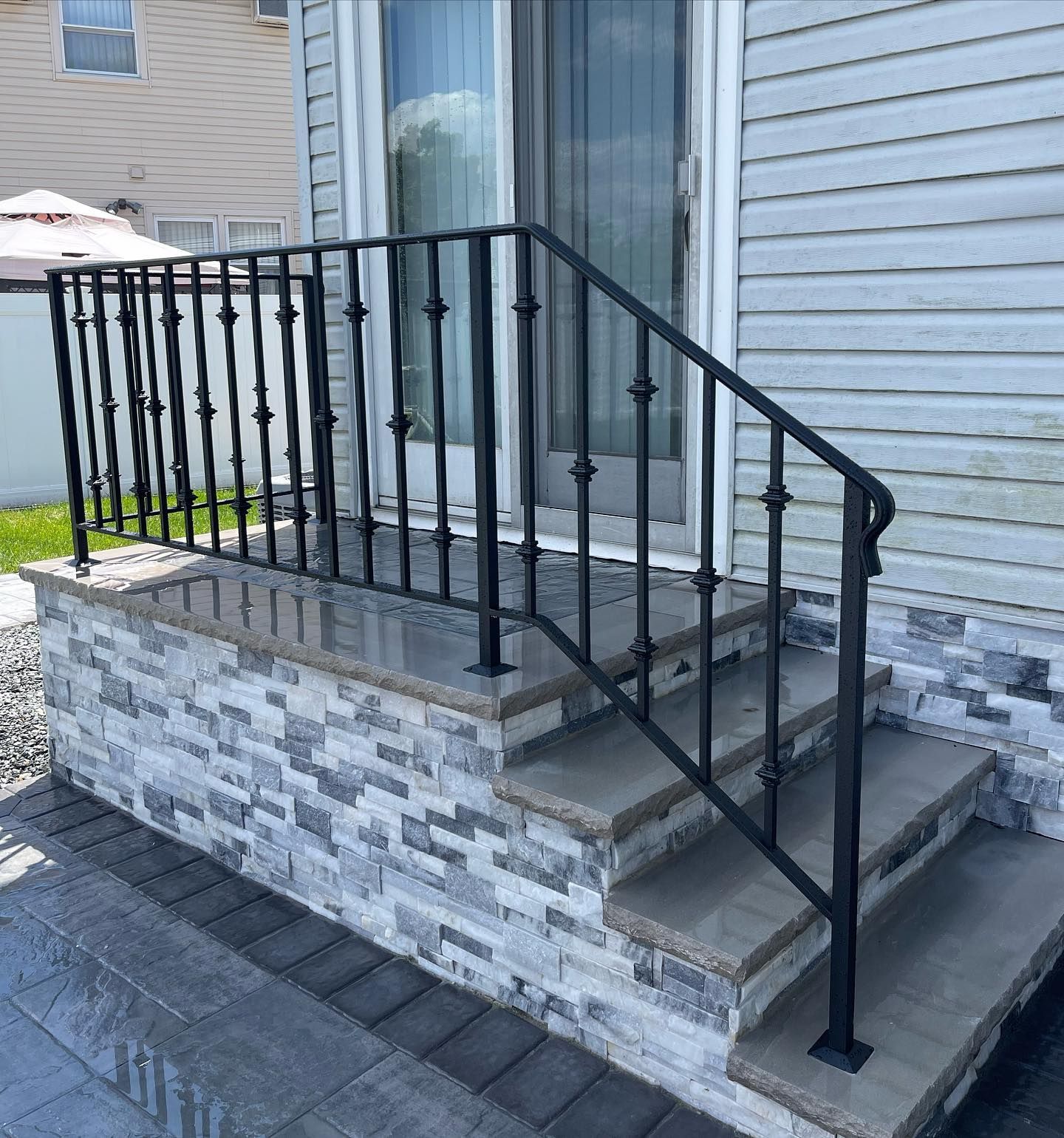 A concrete porch with grey stone veneer, light stone steps, and black metal railings leading to a sliding glass door.
