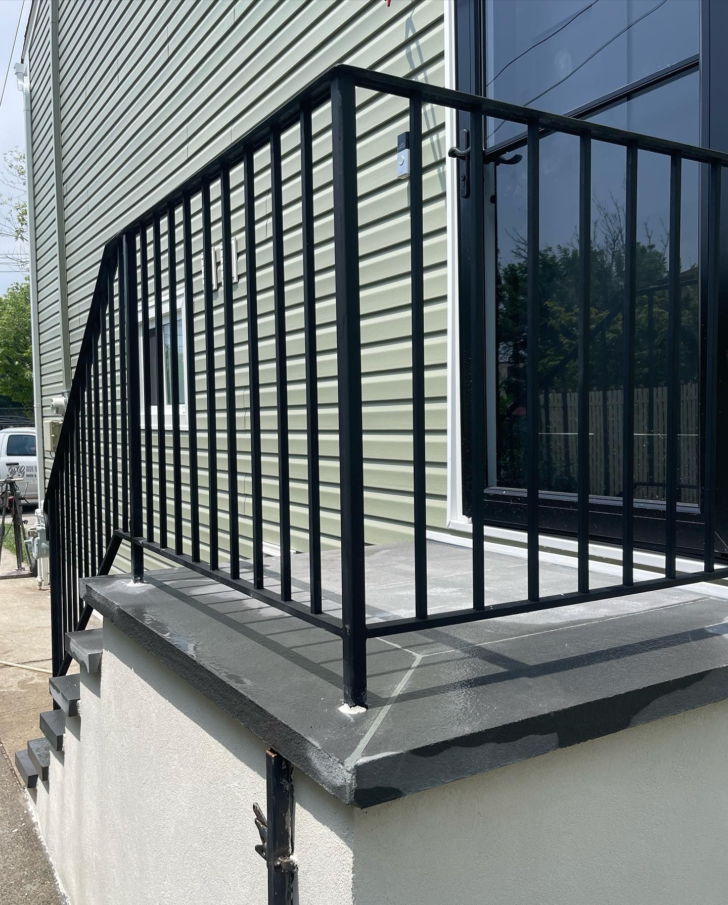 Black metal railing installed along the edge of a concrete porch leading to a building with light green siding.