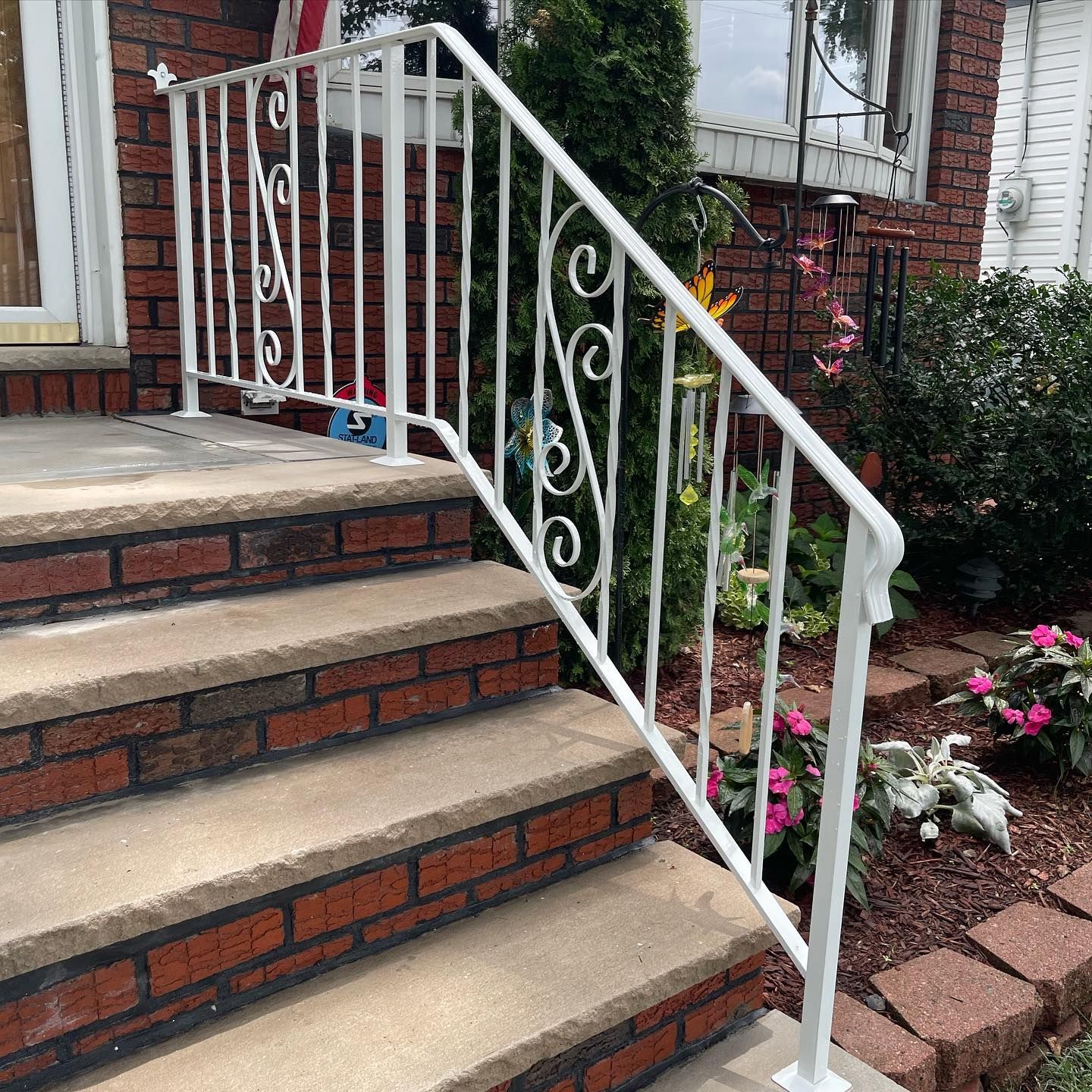 A white, decorative metal handrail runs along stone steps leading to a brick house entrance with garden landscaping.