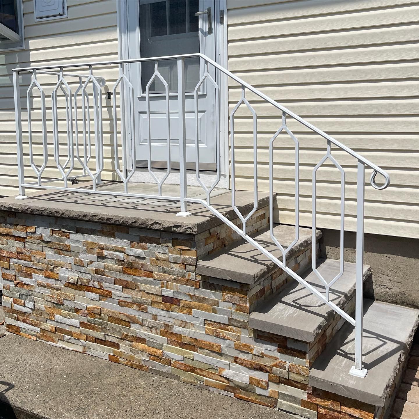Concrete steps with stone-veneer sides and a white metal railing leading to a house door.