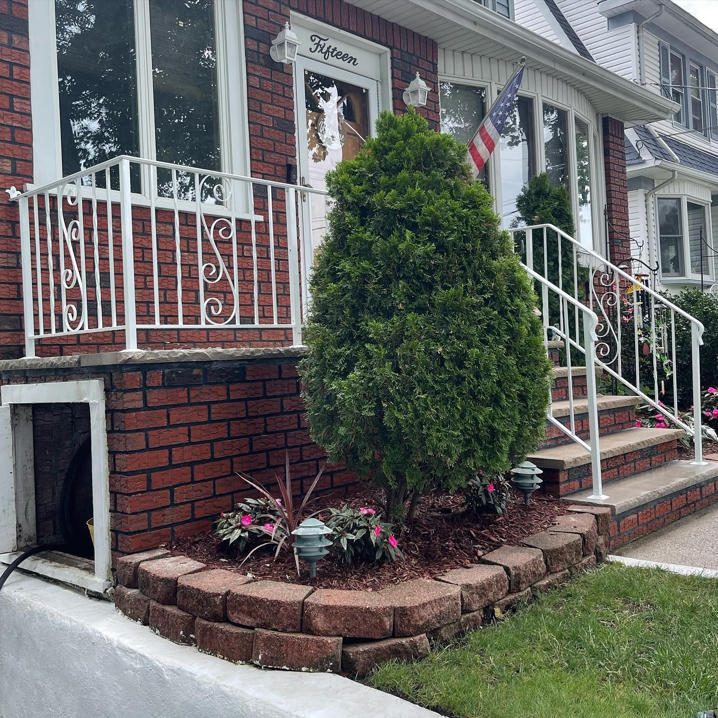 A brick house facade with a white iron porch railing, a tall evergreen, and a tiered garden bed of mulch and flowers.