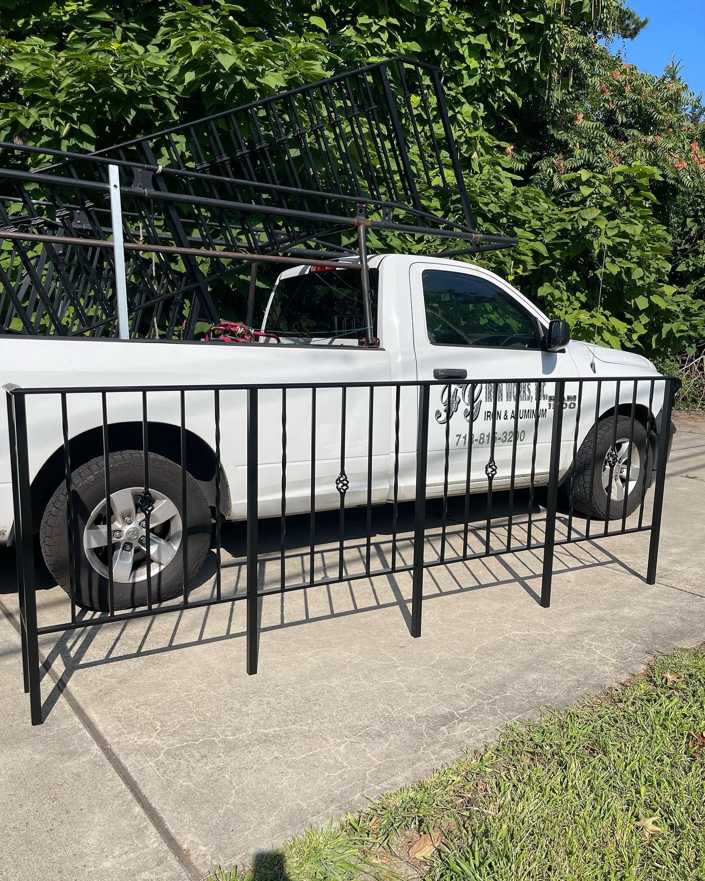 A white pickup truck with a black metal fence section attached to its side, parked on a concrete driveway near trees.