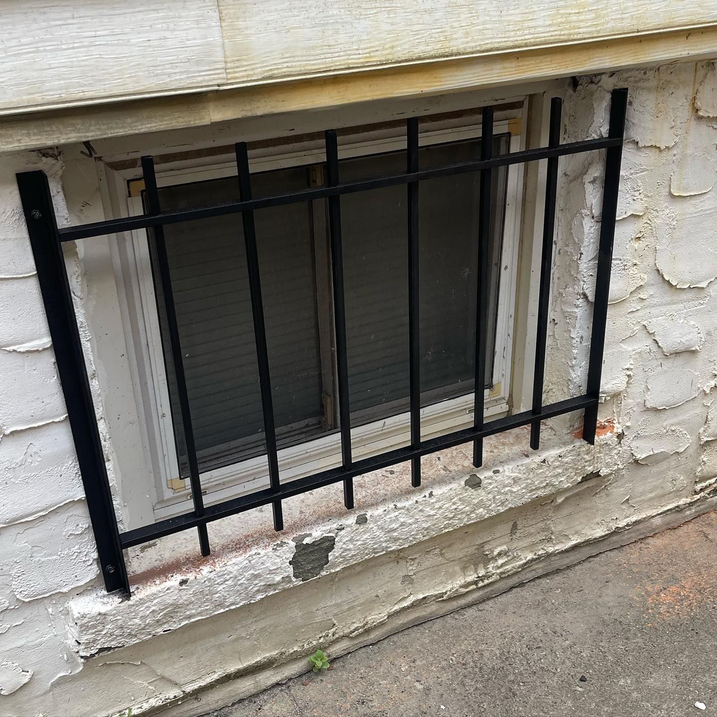 A black metal security grate mounted over a rectangular basement window on a white, weathered stone exterior wall.