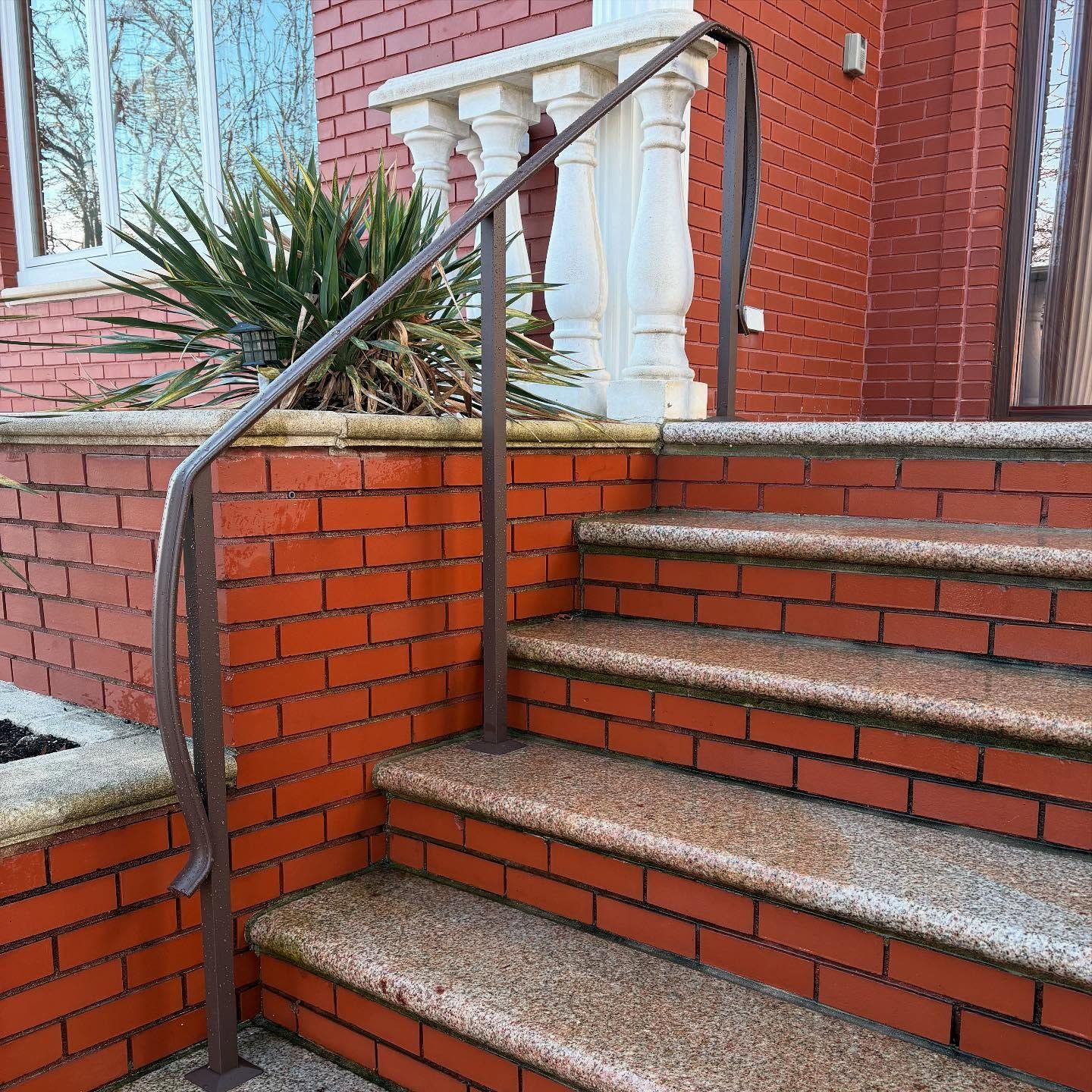 A brown metal handrail extends alongside stone steps leading to a brick building with a decorative white balustrade.
