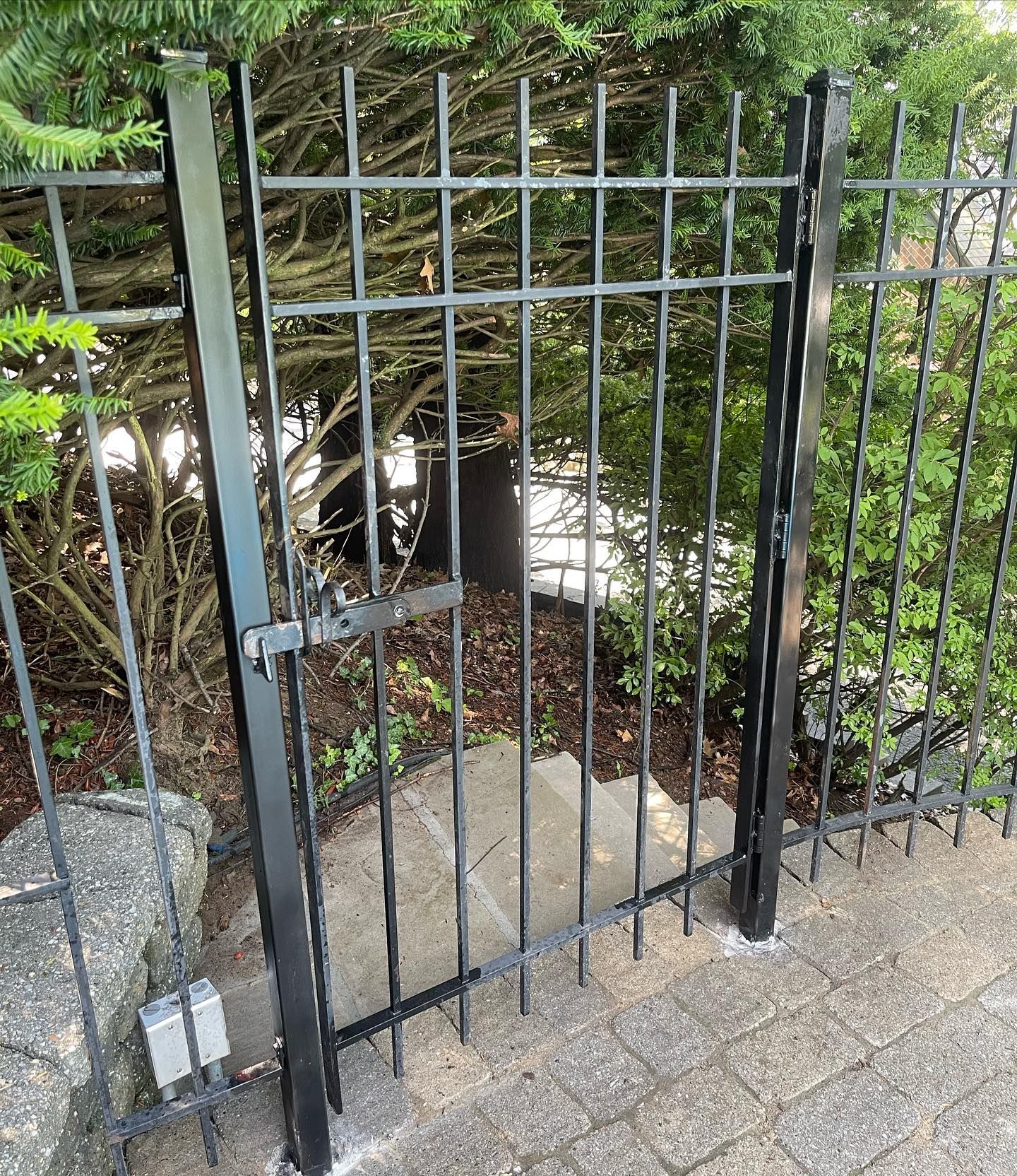 A black metal garden gate in a brick pathway, bordered by green bushes and a stone wall.