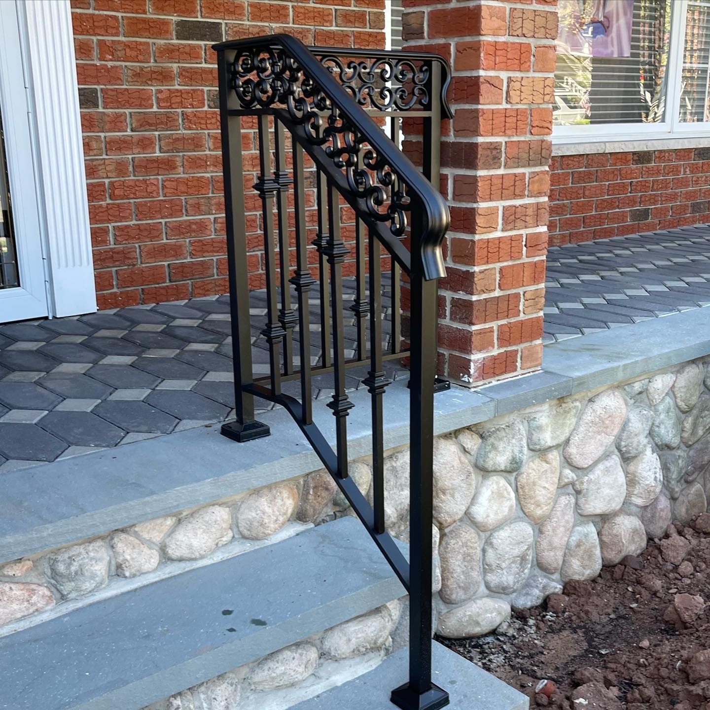 Black ornate metal handrail installed on concrete steps leading to a brick home with a stone-accented porch.