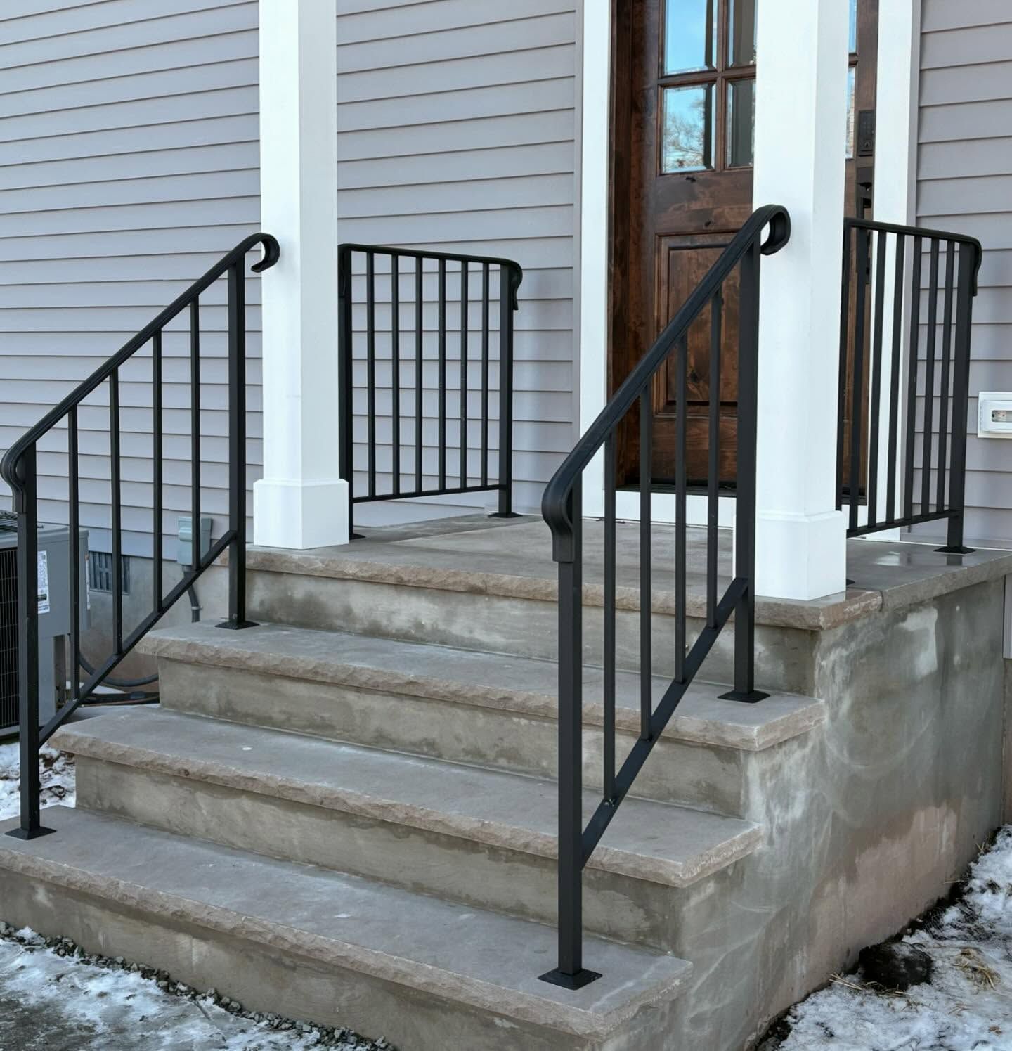 Black metal handrails attached to concrete steps leading up to a house entrance with a wooden door.