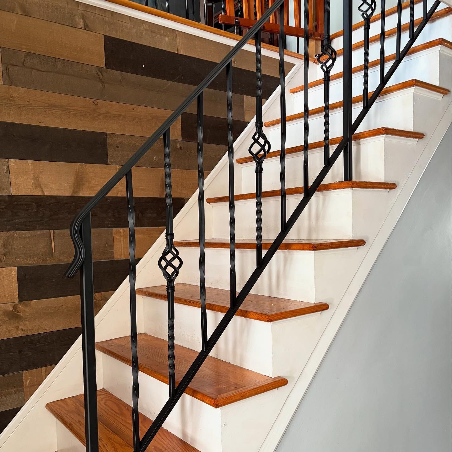 A staircase with wooden treads, white risers, and a decorative black wrought iron railing beside a rustic wood-plank wall.