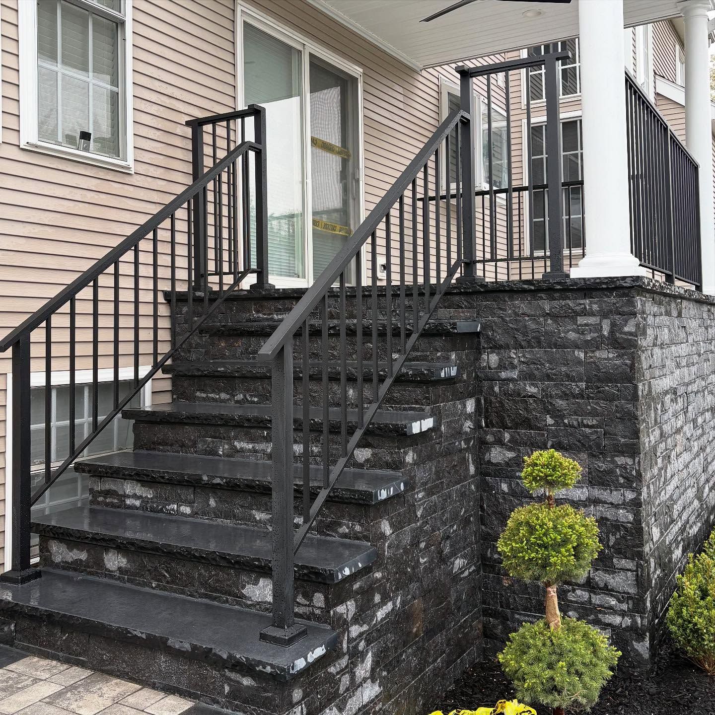 Outdoor stone stairs with black metal railings leading up to a house entrance with light-colored siding.