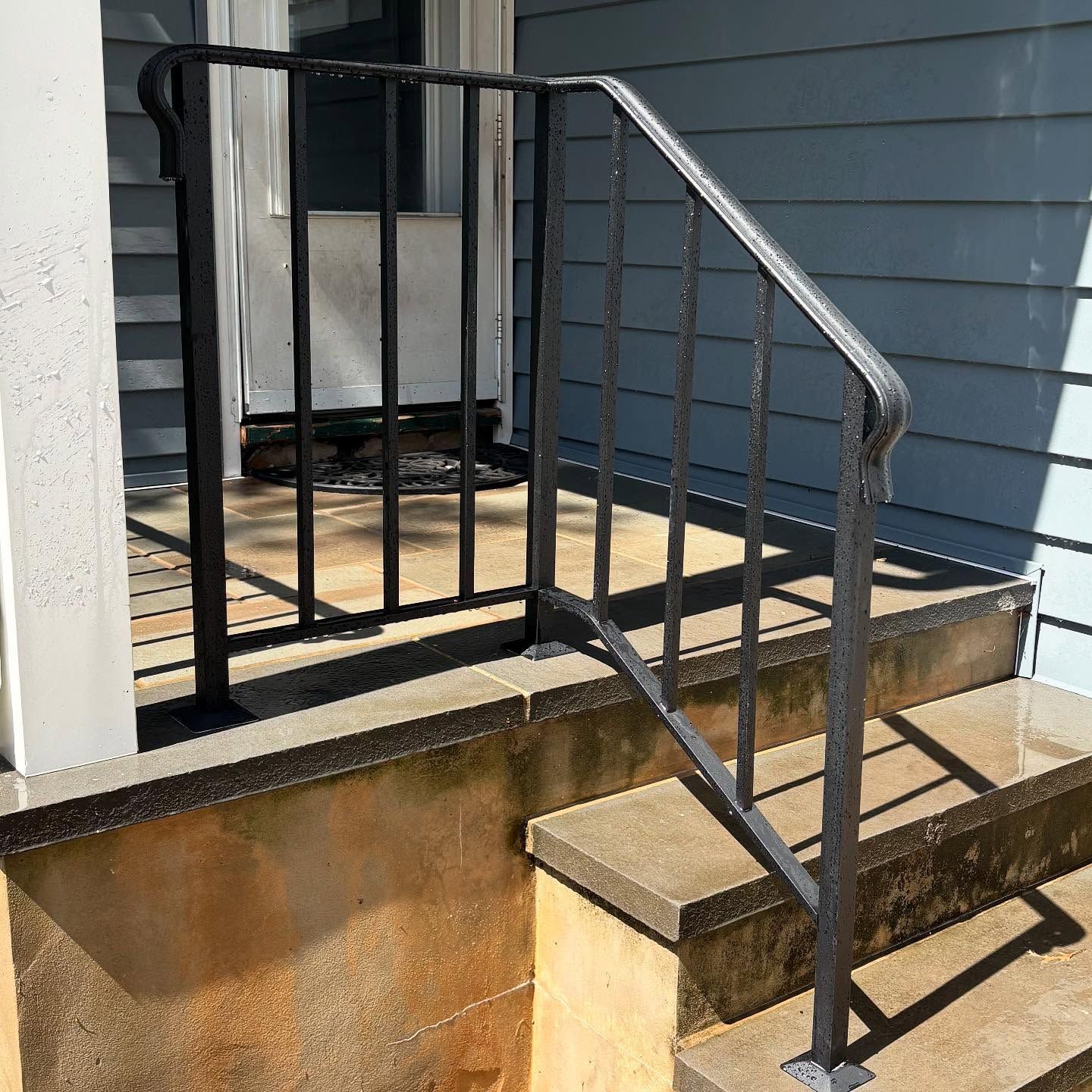 A black metal handrail installed on concrete stairs leading up to a house porch with blue siding.