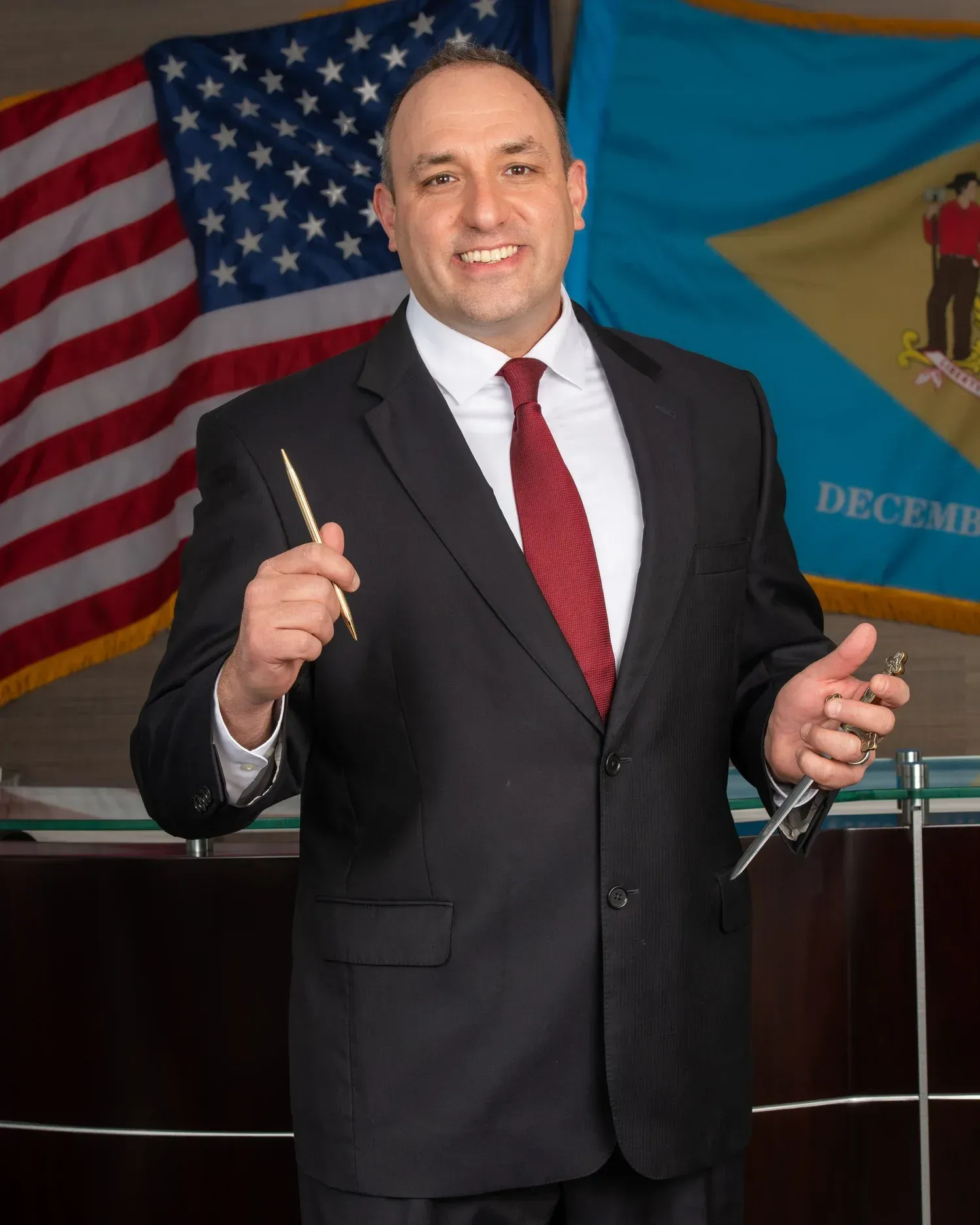 Man in a dark suit and red tie speaking at a podium with U.S. and state flags behind him