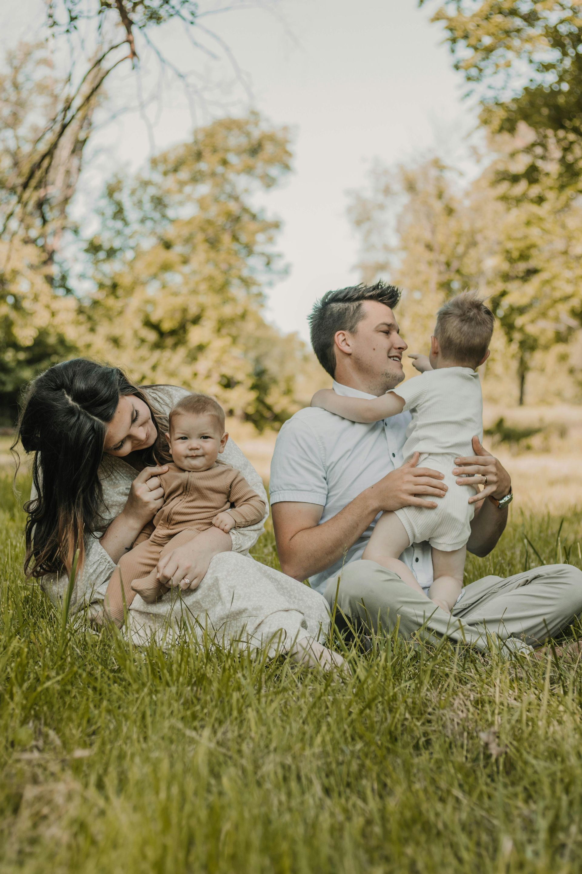 Family sitting in tall grass outdoors, smiling and holding two small children in a sunny autumn park
