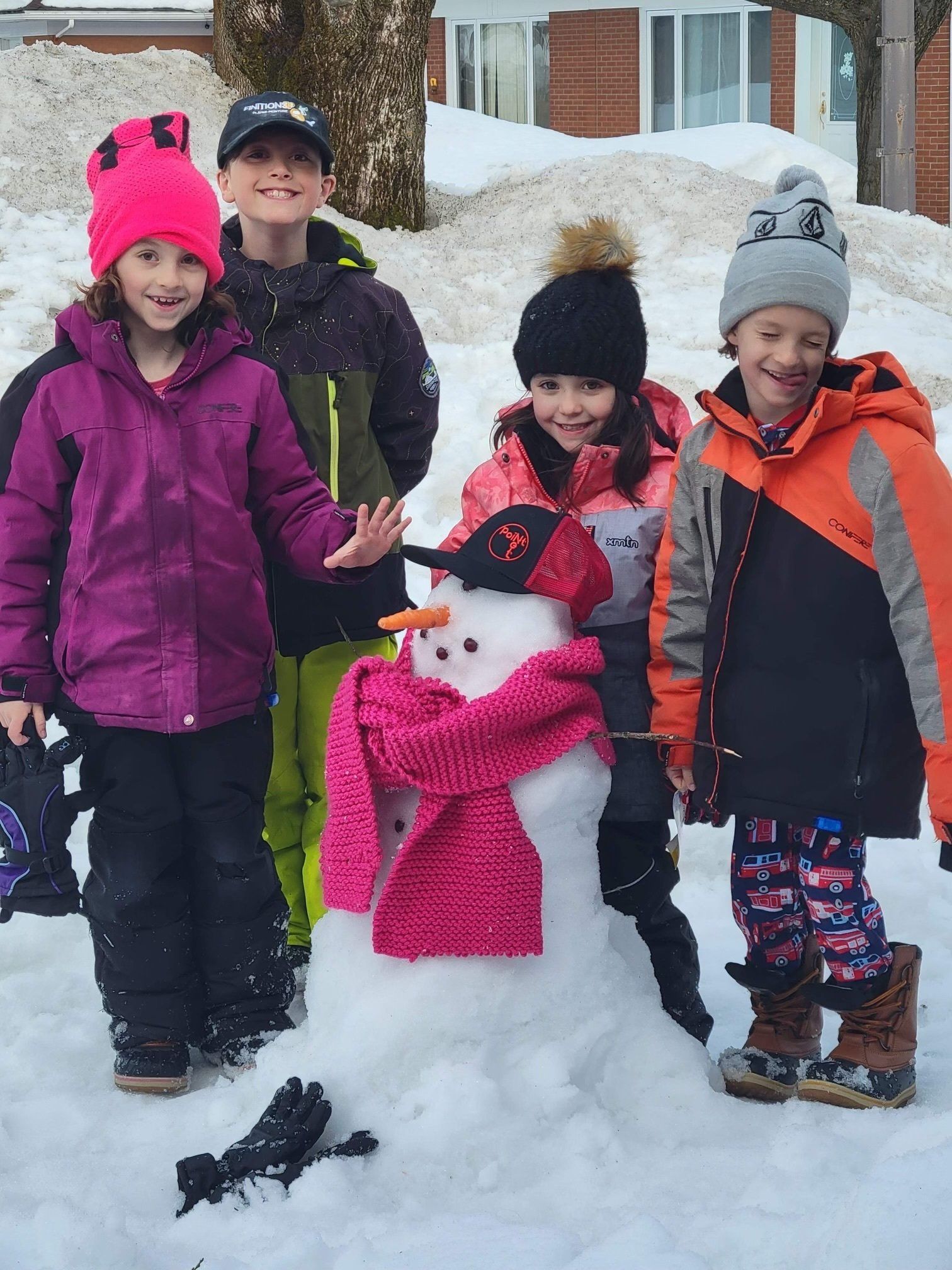 Des enfants posent avec un bonhomme de neige portant des accessoires roses dans un décor enneigé.