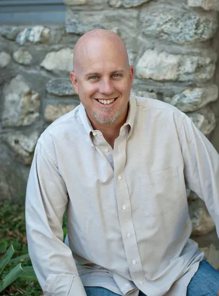 A bald man in a tan shirt is smiling in front of a stone wall