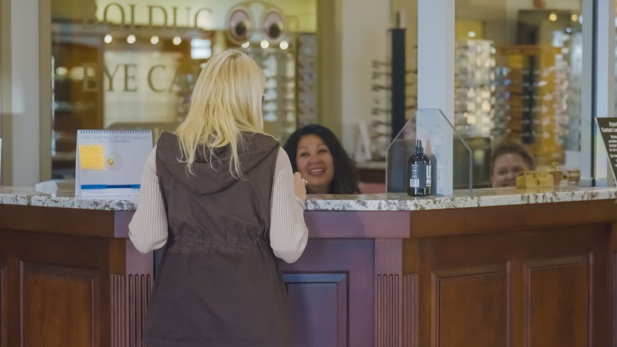 Woman stands at a café counter, speaking with two smiling staff members behind the register.