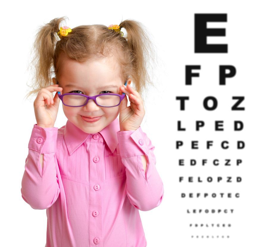 A little girl wearing glasses is standing in front of an eye test chart.