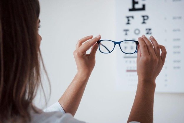 A woman is holding a pair of glasses in front of an eye chart.