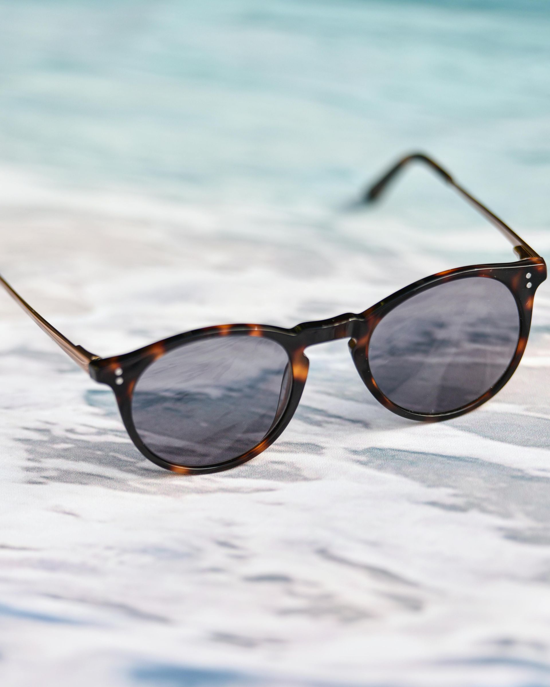 Tortoiseshell sunglasses on a sandy beach with blurred ocean background.