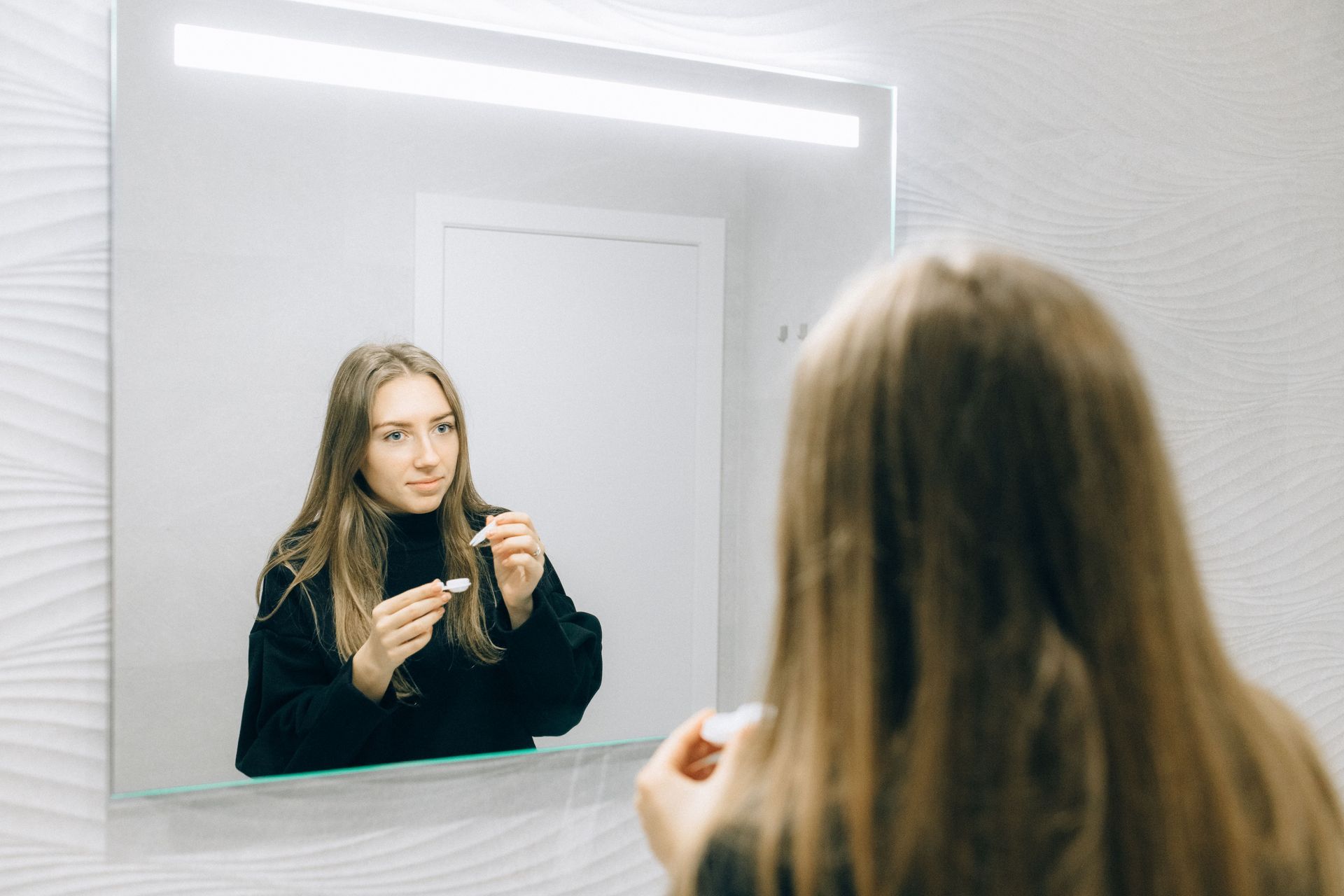 Person applying makeup in a bathroom mirror, holding a lipstick, with a bright backlit vanity mirror