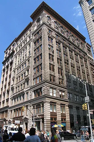 Tall historic brick building on a city street under a blue sky, with pedestrians and cars below.