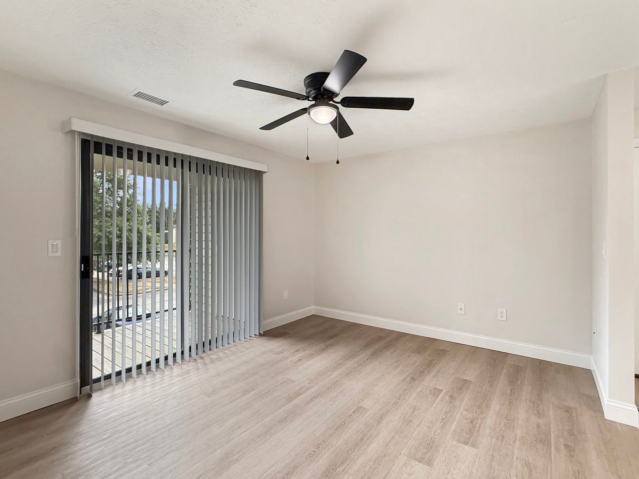 Empty modern living room with light wood floors, ceiling fan, and sliding glass door with vertical blinds