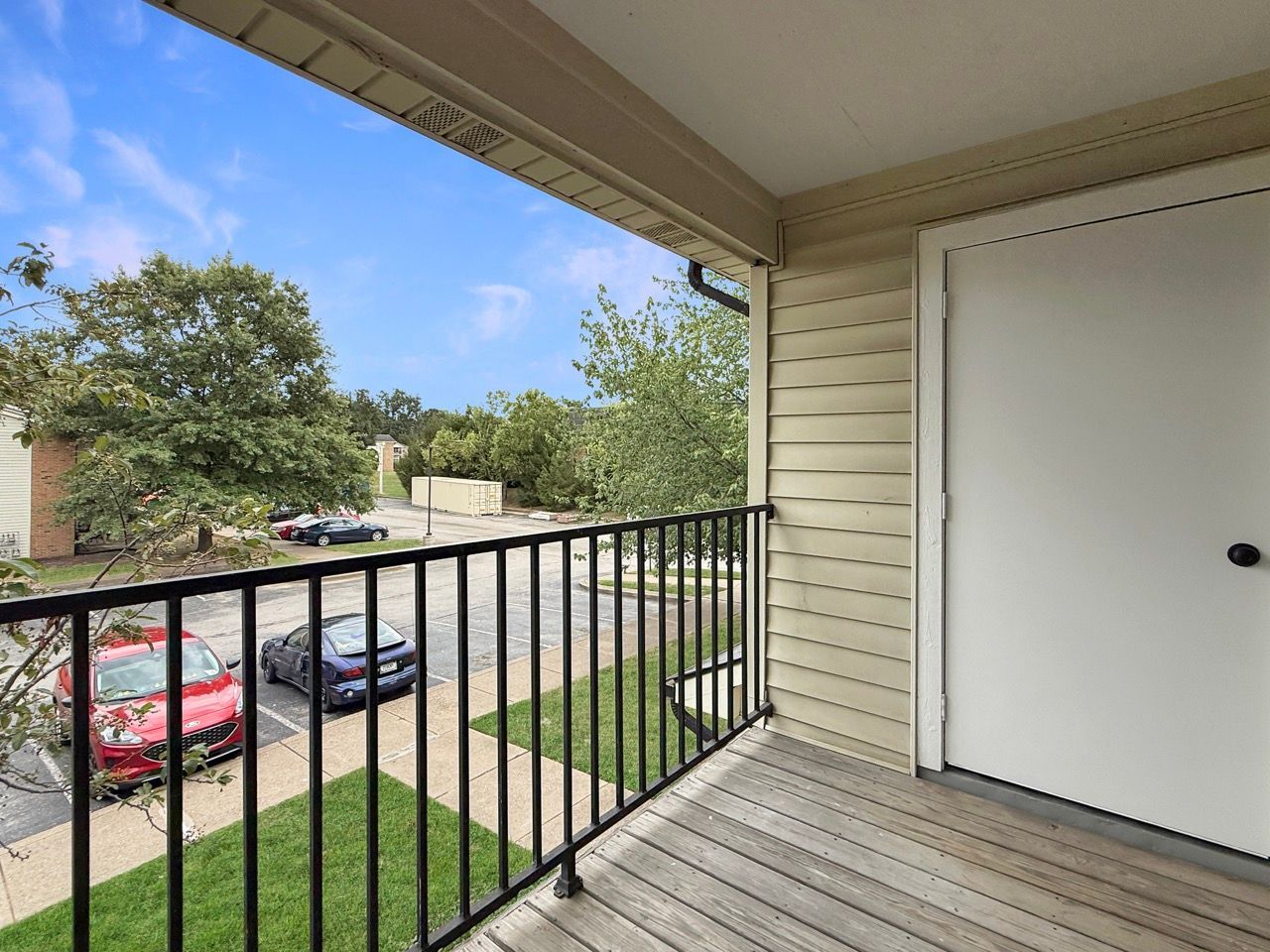 Apartment balcony overlooking a parking lot and trees, with black railing and white door.