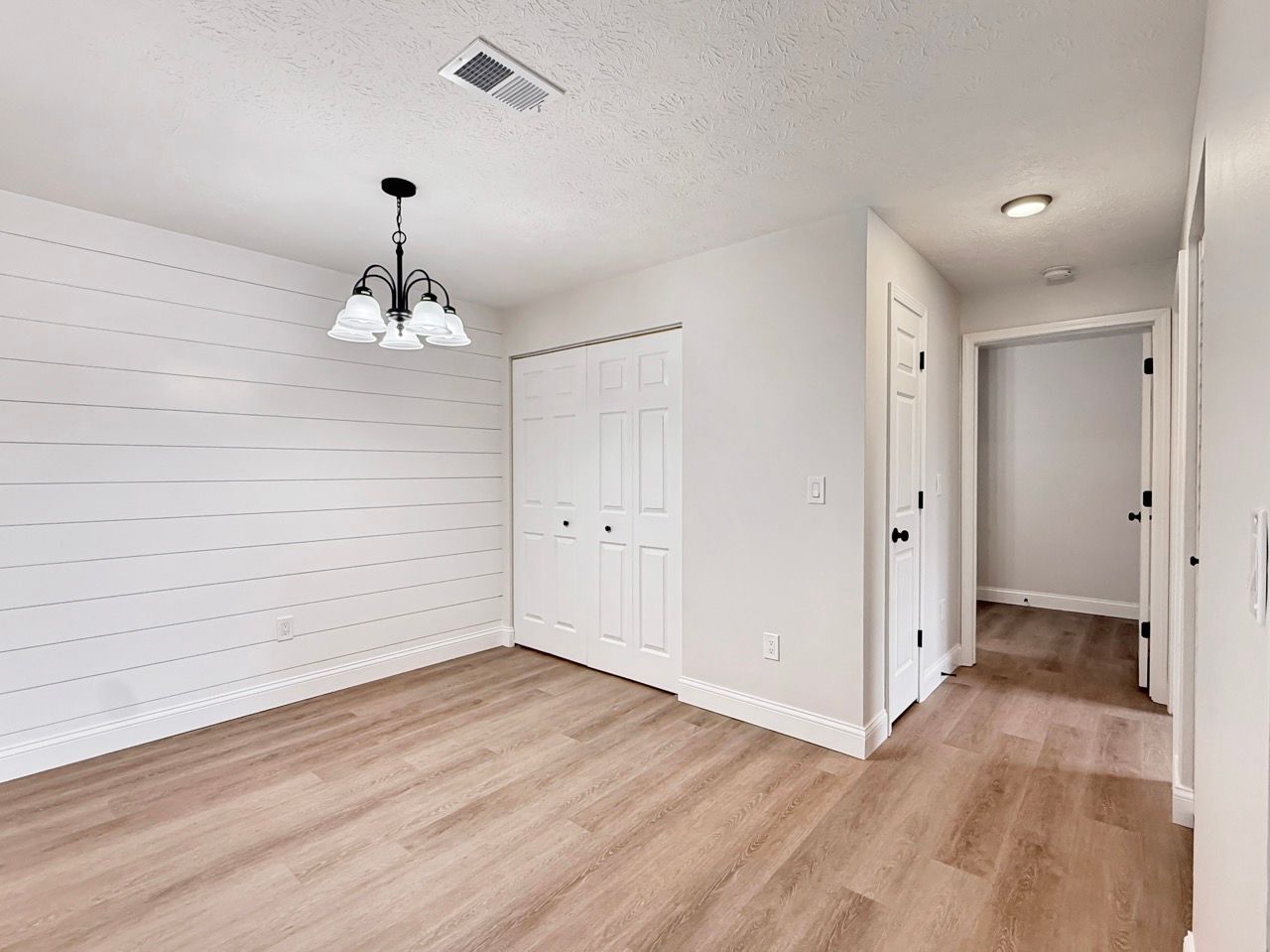 Empty white room with wood floors, ceiling light, closet doors, and hallway entry.