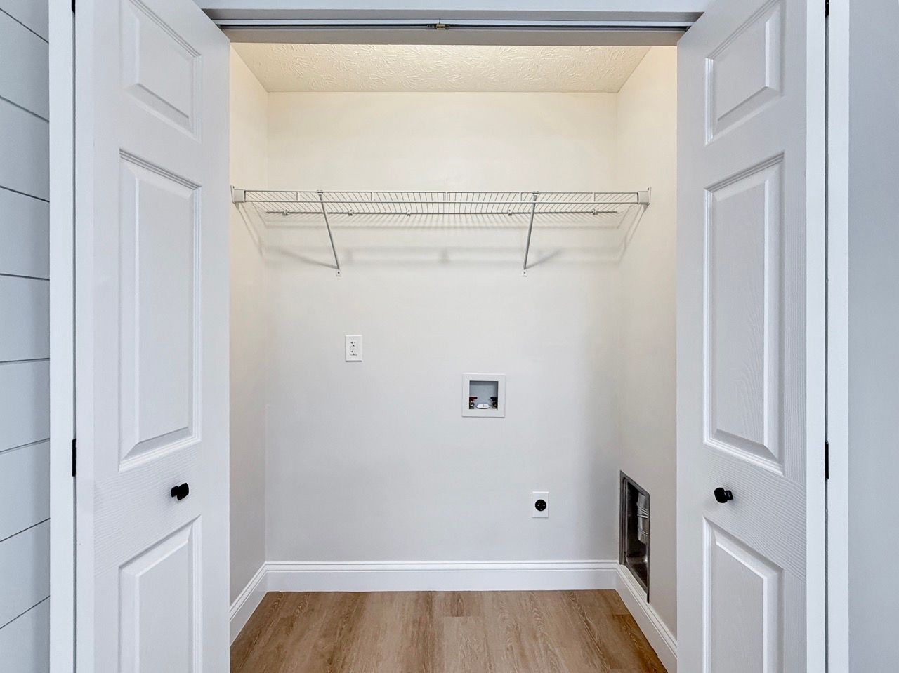 Empty white laundry closet with wire shelf and wood floor, viewed through open double doors