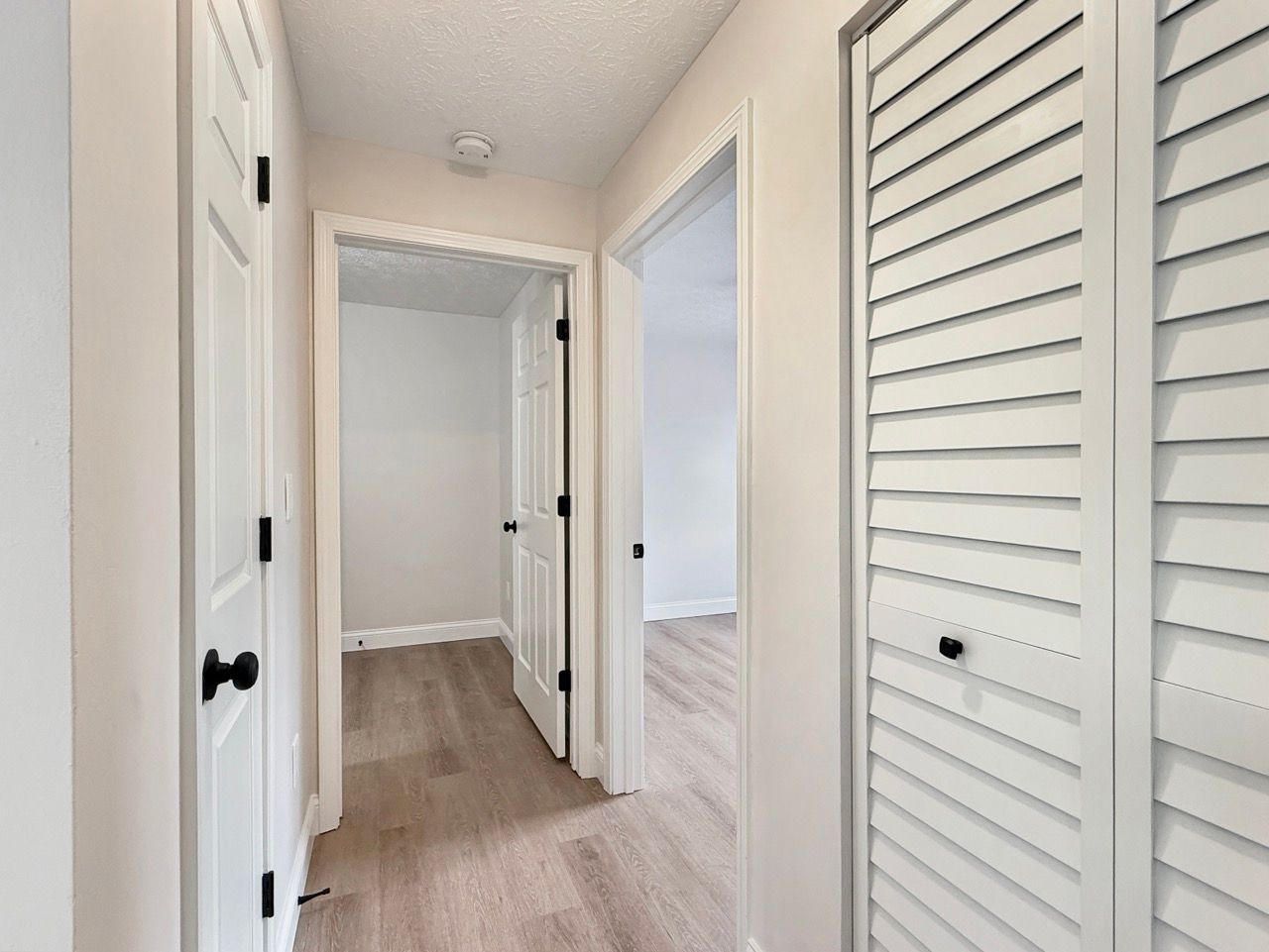 Bright hallway with white walls, louvered closet doors, and light wood flooring leading to a bedroom.