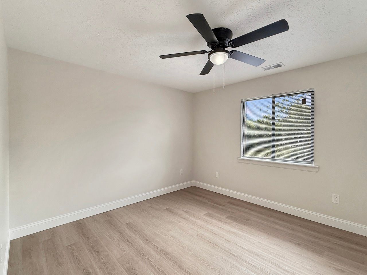 Empty beige bedroom with a dark ceiling fan, light wood floor, and a small window showing palm leaves.