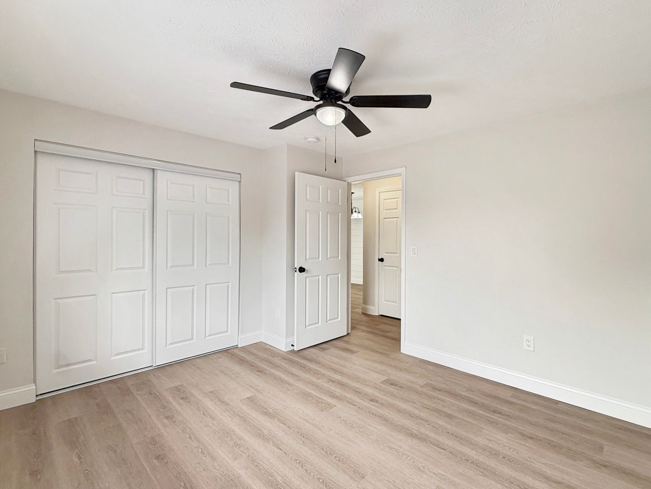 Empty bedroom with white closets, light wood flooring, and a black ceiling fan.