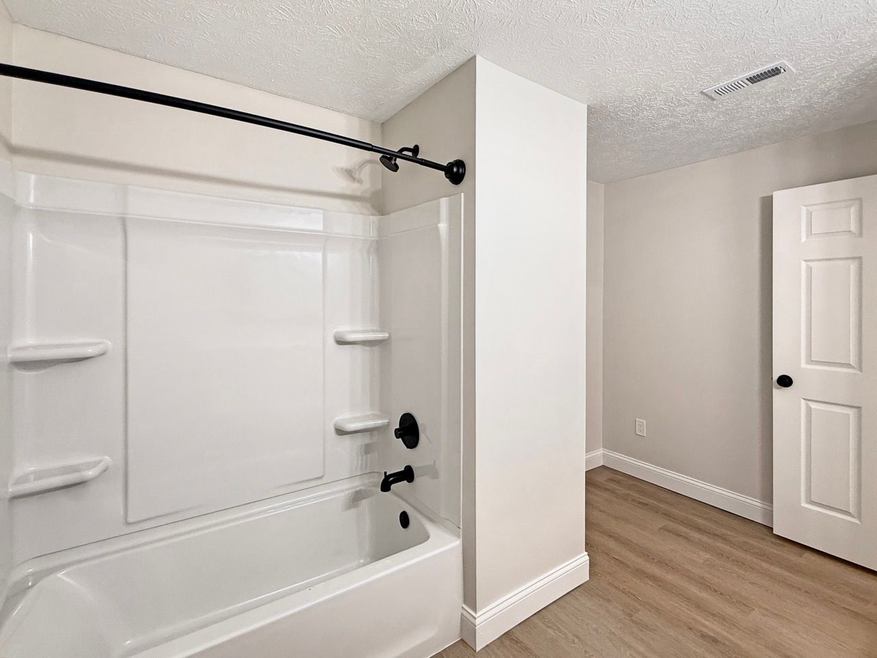 Bright bathroom with white tub, tiled shower surround, and wood-look flooring beside a white door.