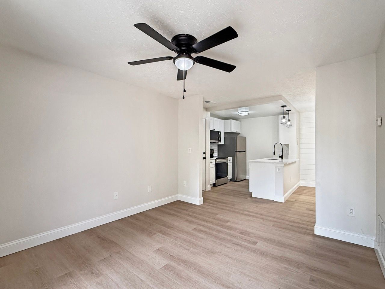 Empty white living room with ceiling fan, wood floors, and open view into a bright kitchen.