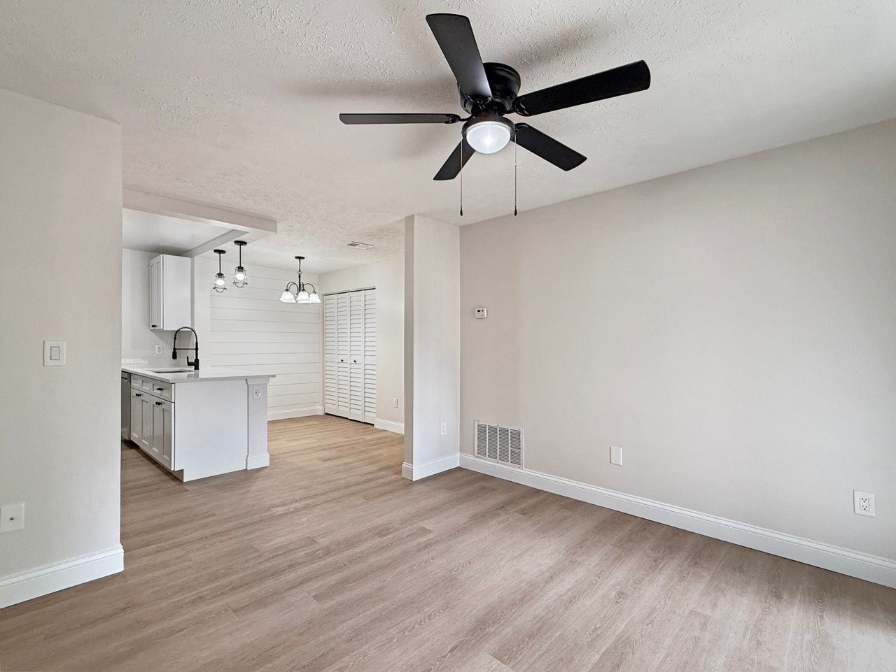 Empty living room with wood floors, white walls, ceiling fan, and open view to kitchen and hallway.