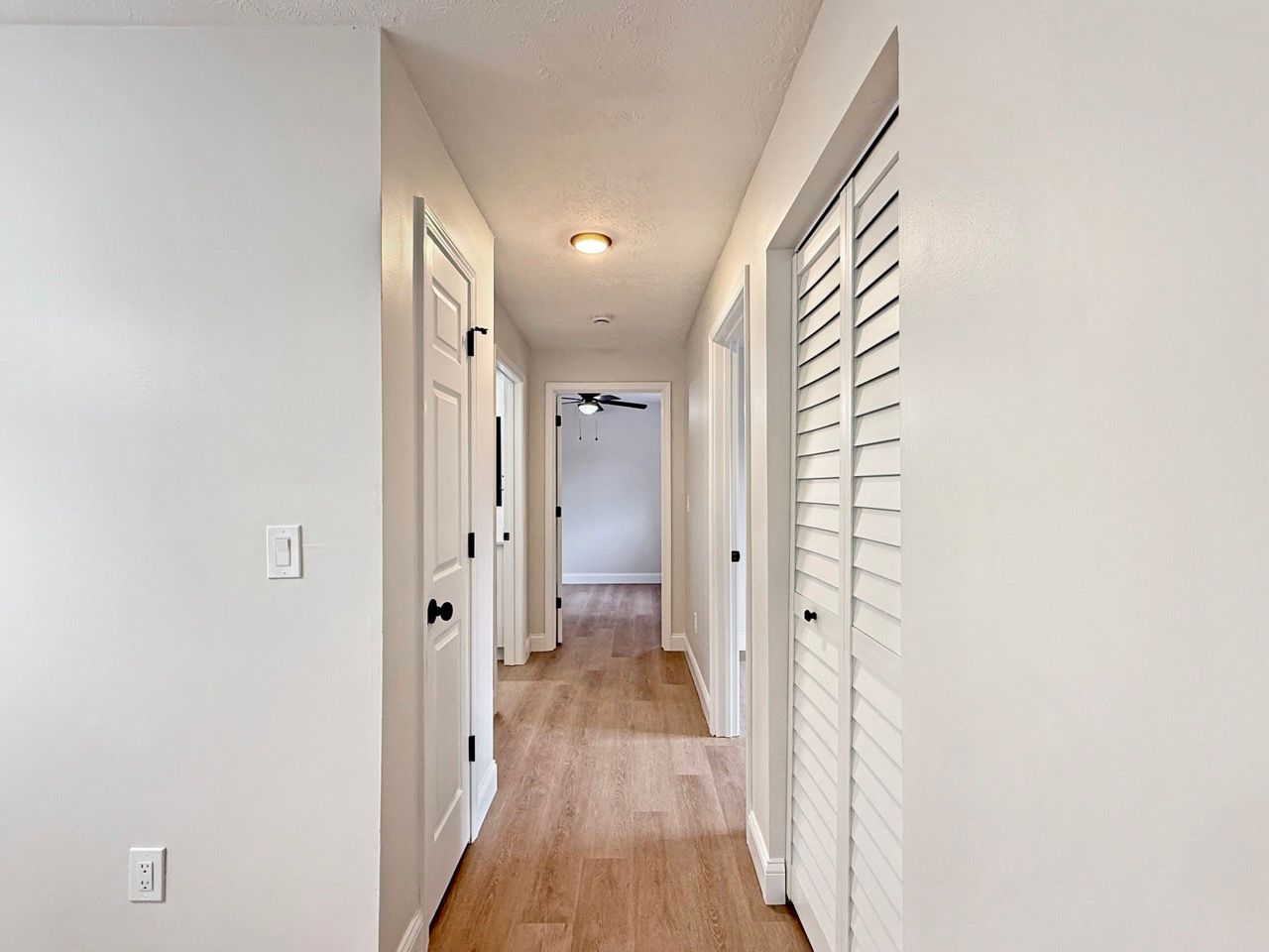 Narrow white hallway with wood floor, closet doors, and a ceiling light.