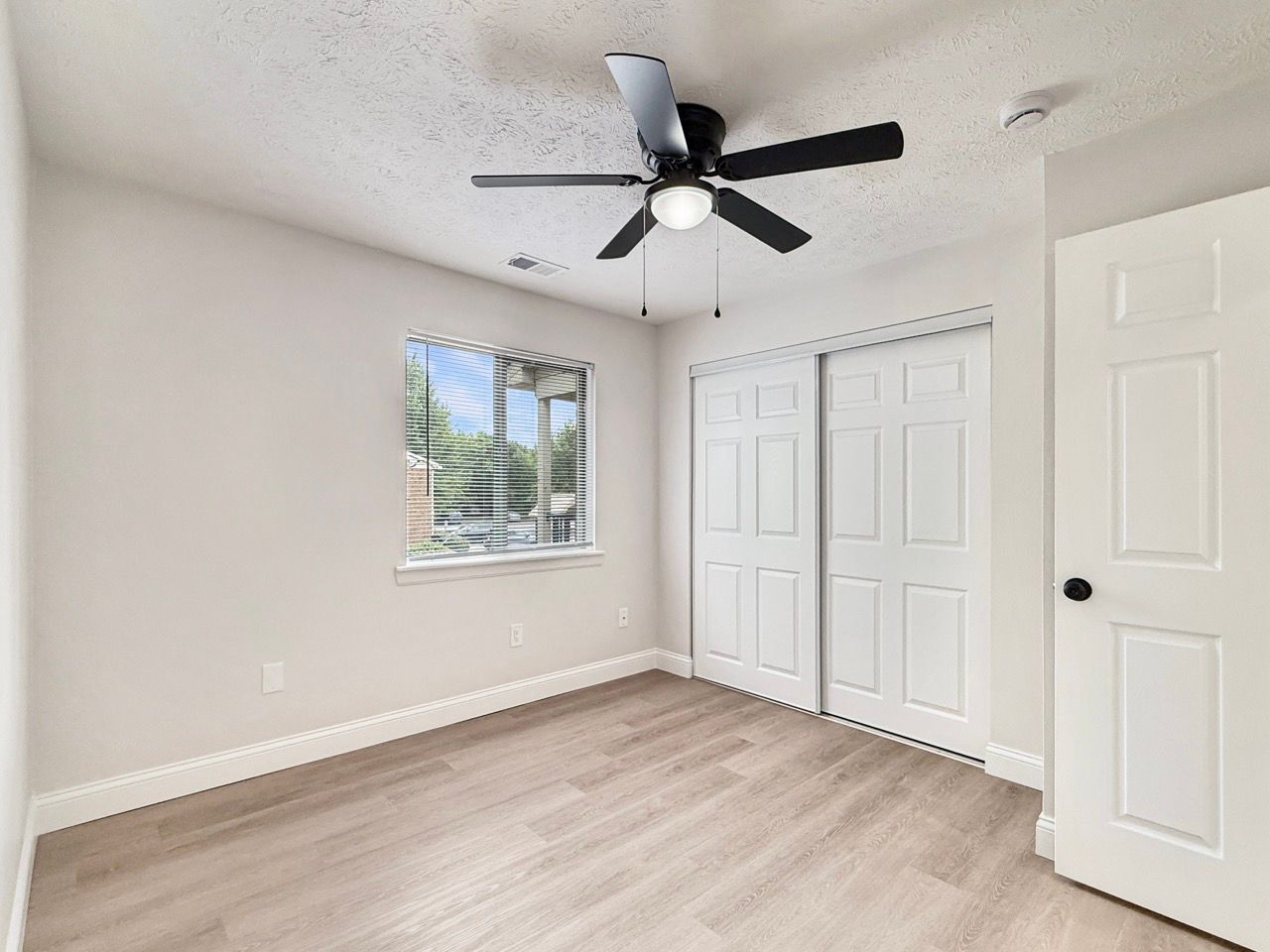 Empty beige bedroom with white closet doors, ceiling fan, and window light through a small window.