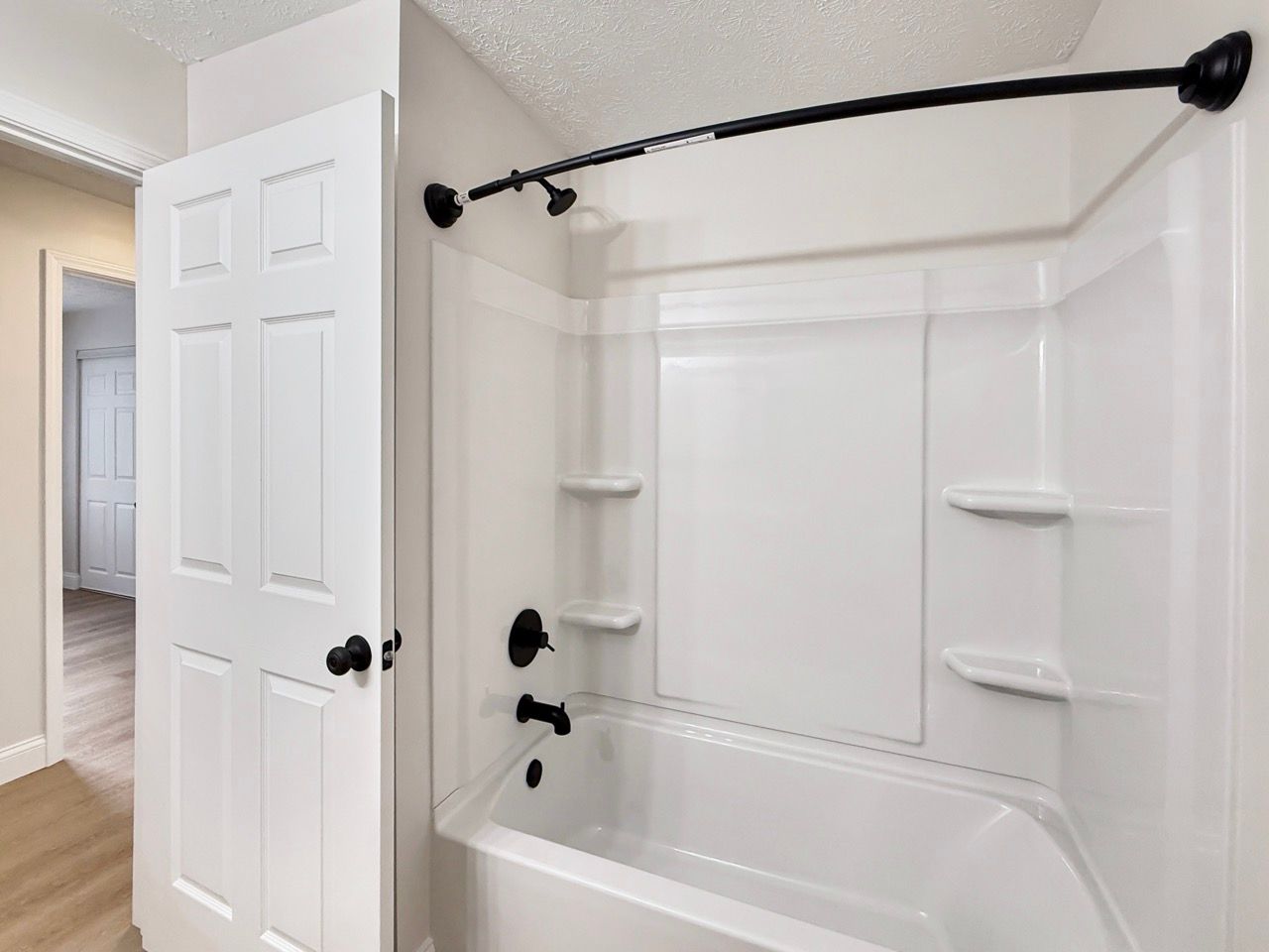 Bathroom with white tub-shower, black curtain rod, built-in shelves, and an open door.
