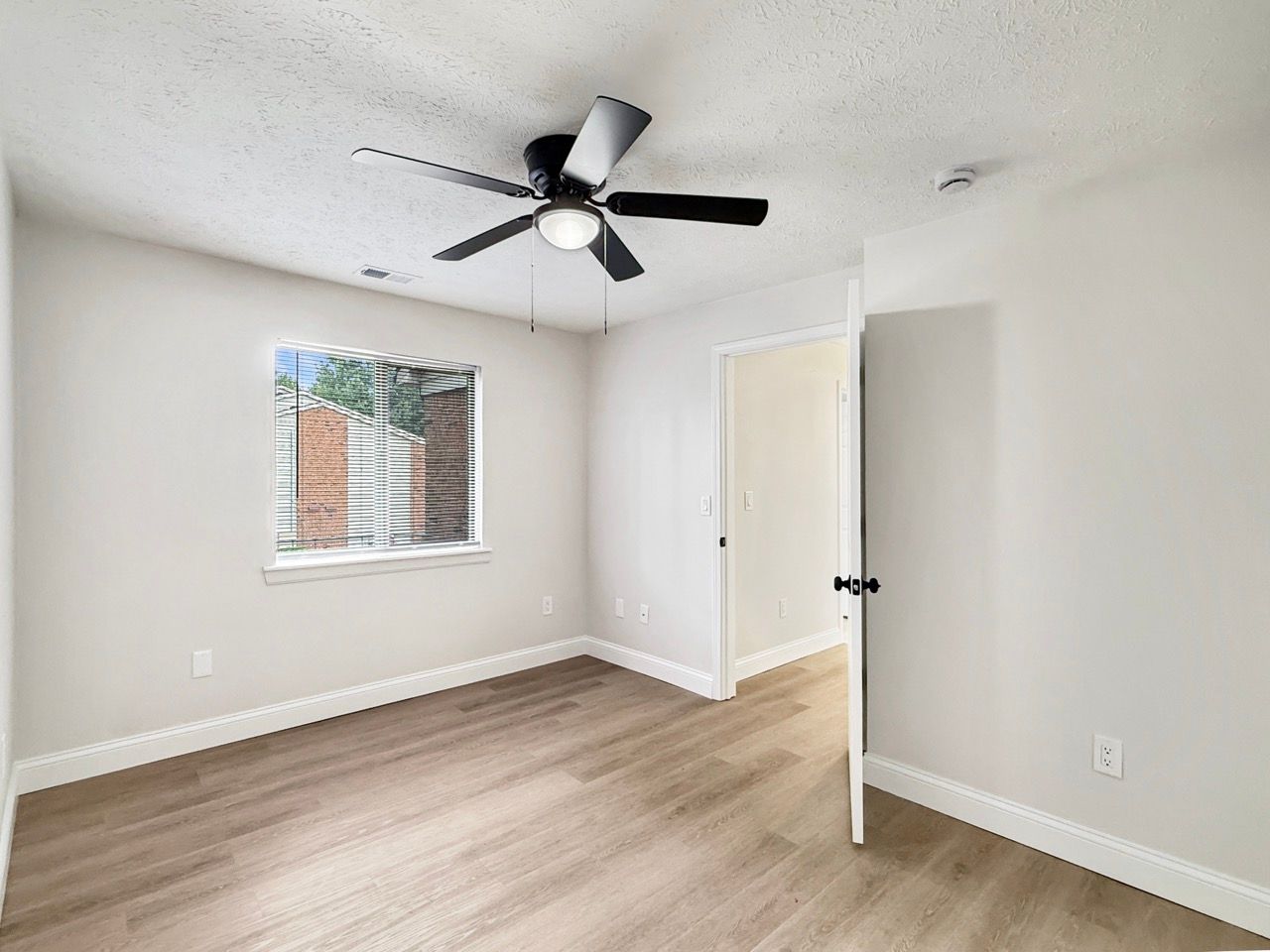 Empty white bedroom with light wood floors, ceiling fan, and an open door near a window.